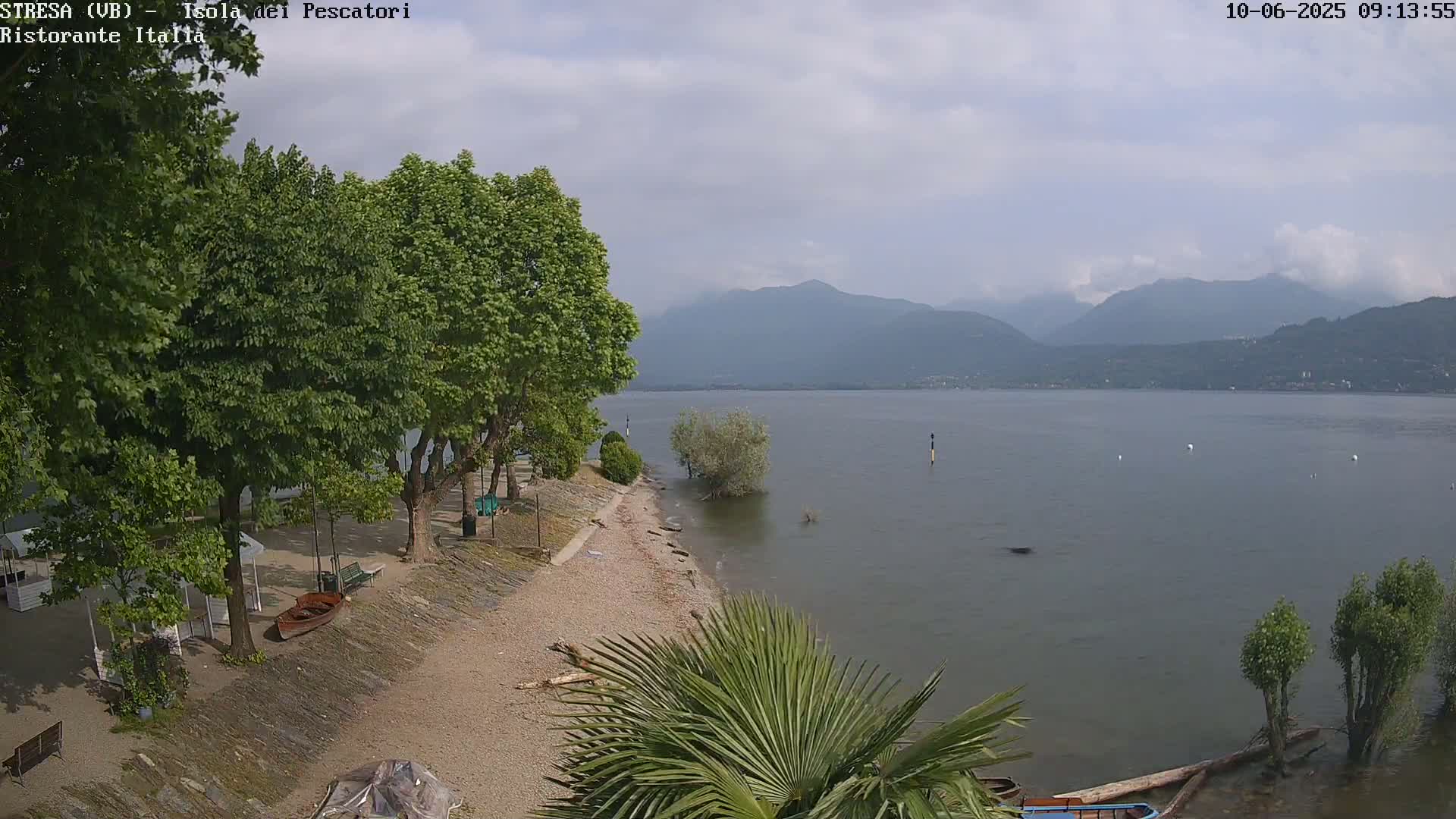 A partly cloudy day reveals a lakeside view with a sandy beach, several trees, small boats, and distant mountains.