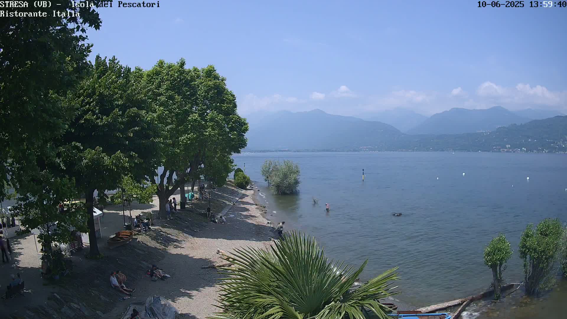 A sunny day at a lakeside beach with people relaxing under trees, mountains in the distance, and calm water.