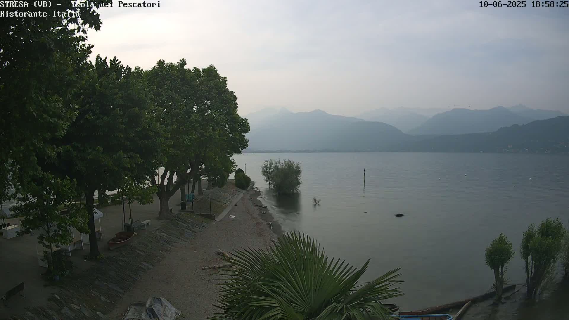 A hazy view of a lake with mountains in the background, seen from a shoreline with trees, a gravel beach, and some small boats.