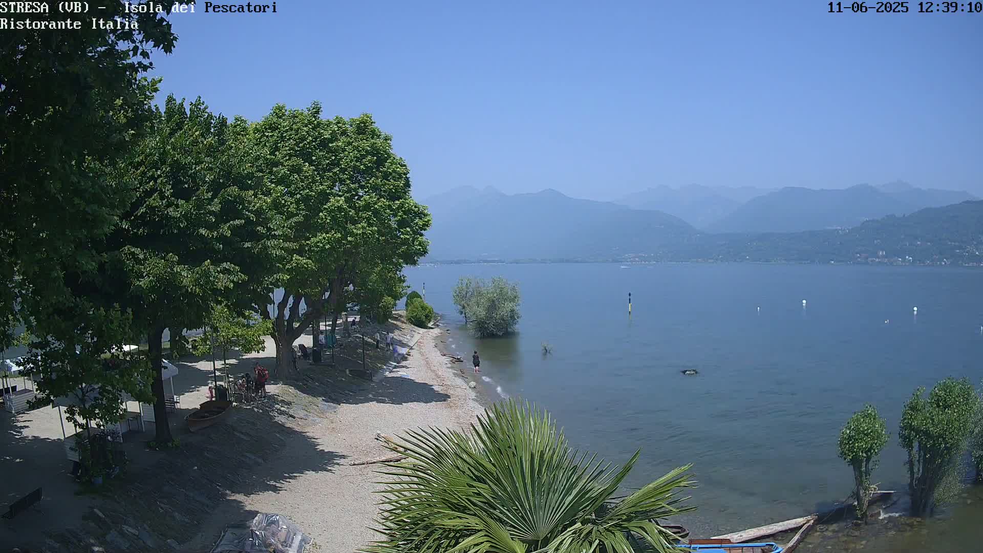 A sandy lakeshore with trees, a few people, and small structures is shown under a clear blue sky, with hazy mountains visible in the distance.