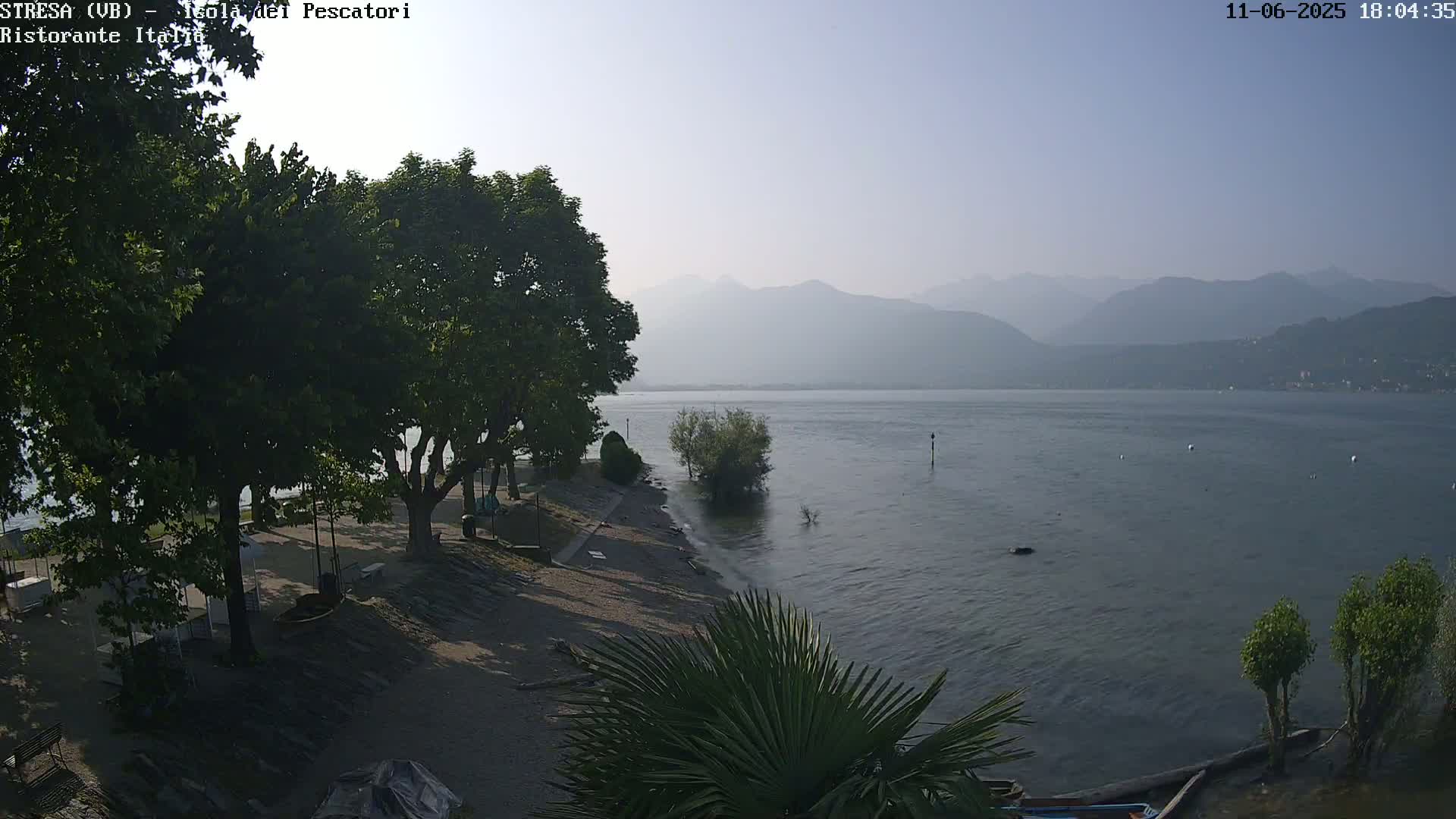 A hazy, sunny day overlooking a calm lake with mountains in the distance, a sandy shore with trees and shrubs in the foreground.