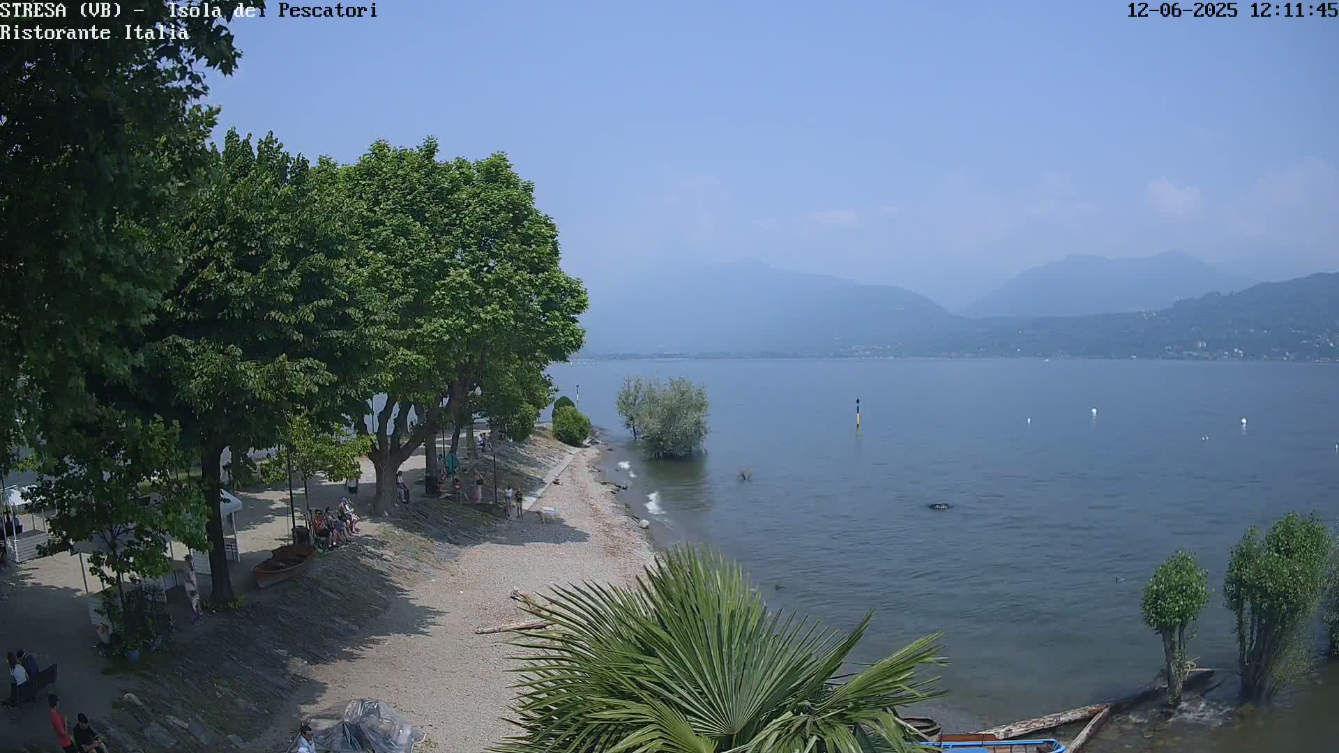 A sunny day at a lakeside beach with people relaxing under trees, a calm lake, and mountains in the distance.