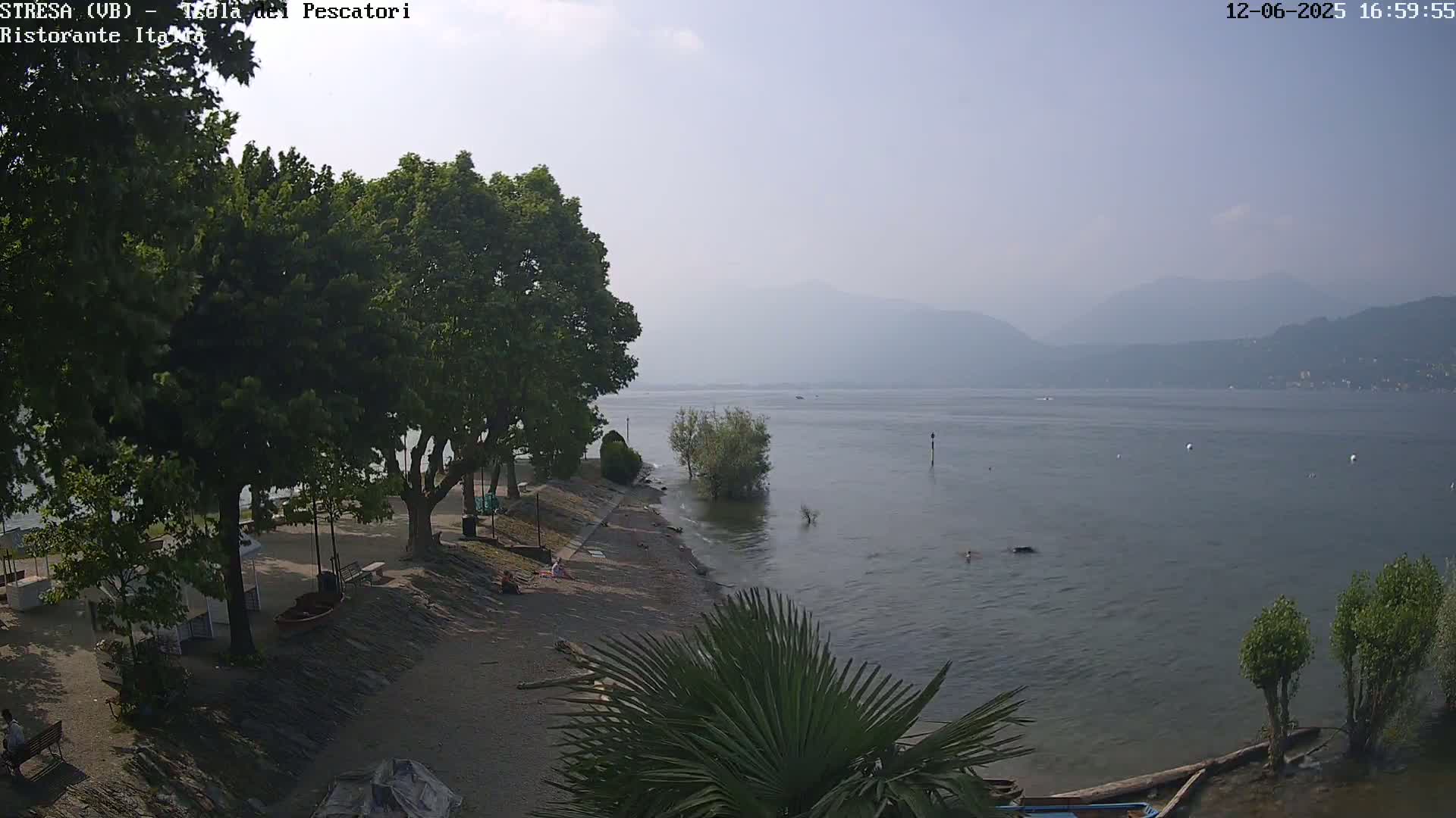 A hazy, sunny day overlooks a calm lake with a rocky shoreline lined with trees, and a few people are visible on the beach.