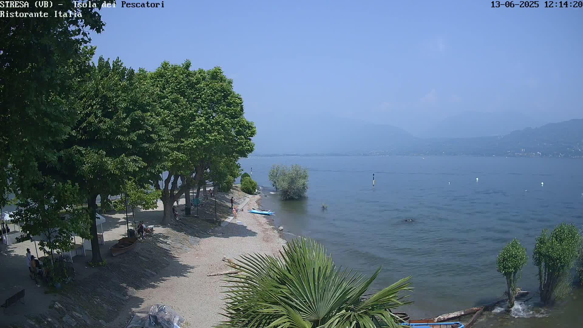 A sandy beach with people and trees lines a calm lake under a clear blue sky on a sunny day, with distant mountains visible in the background.