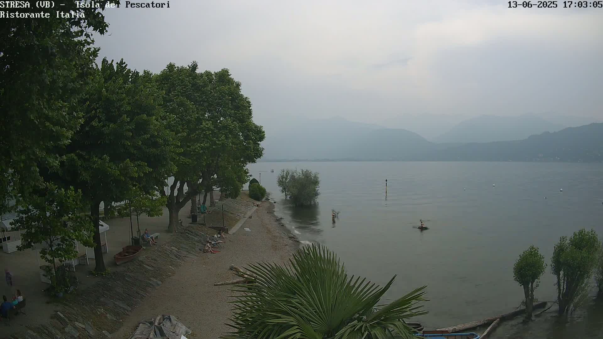 A hazy, overcast day reveals a pebble beach with people relaxing under shady trees, overlooking a calm lake and distant mountains.