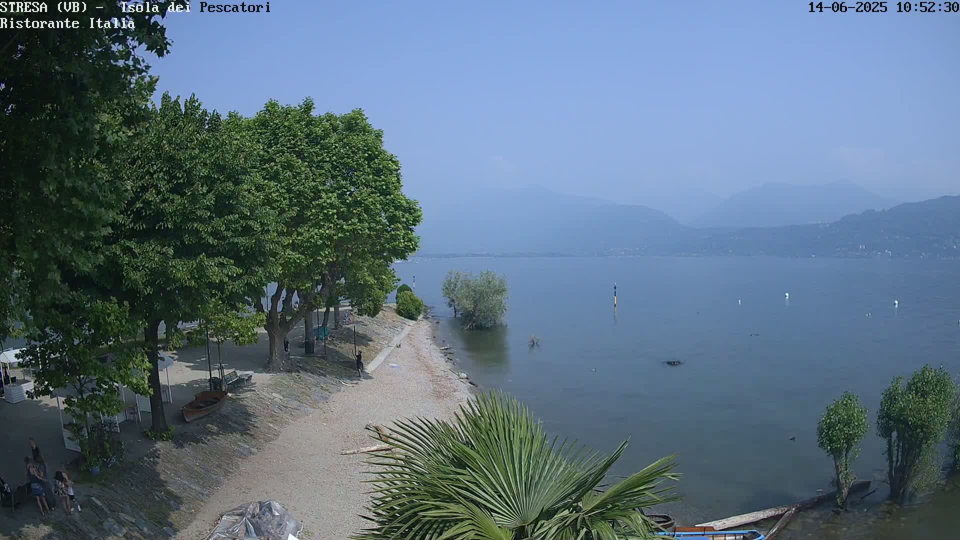 A sandy lakeshore with lush green trees, some people, and a calm lake with hazy mountains in the background under a clear blue sky.