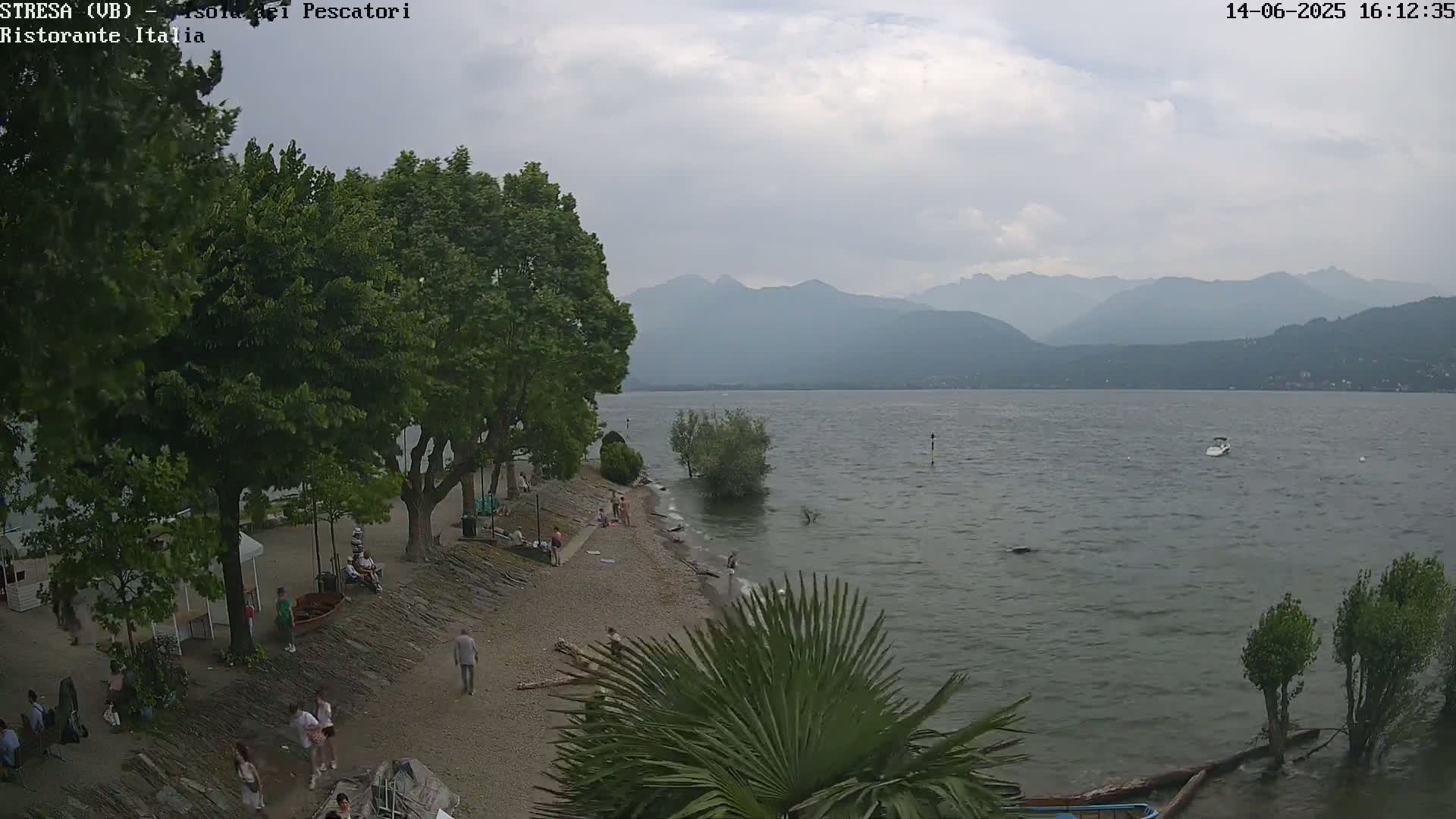 A mostly cloudy day at a lakeside beach with people relaxing and swimming, mountains in the distance.
