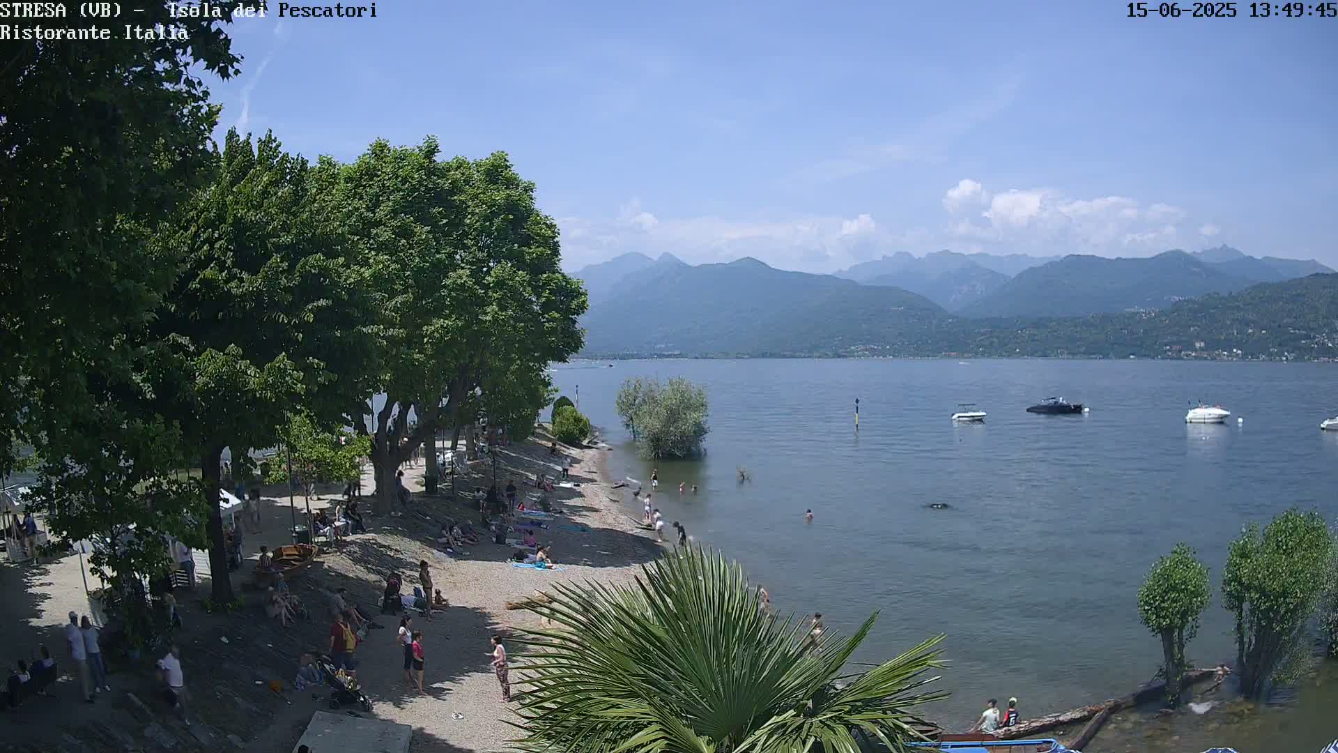 A sunny day at a lakeside beach with people sunbathing and swimming, boats on the water, and mountains in the background.