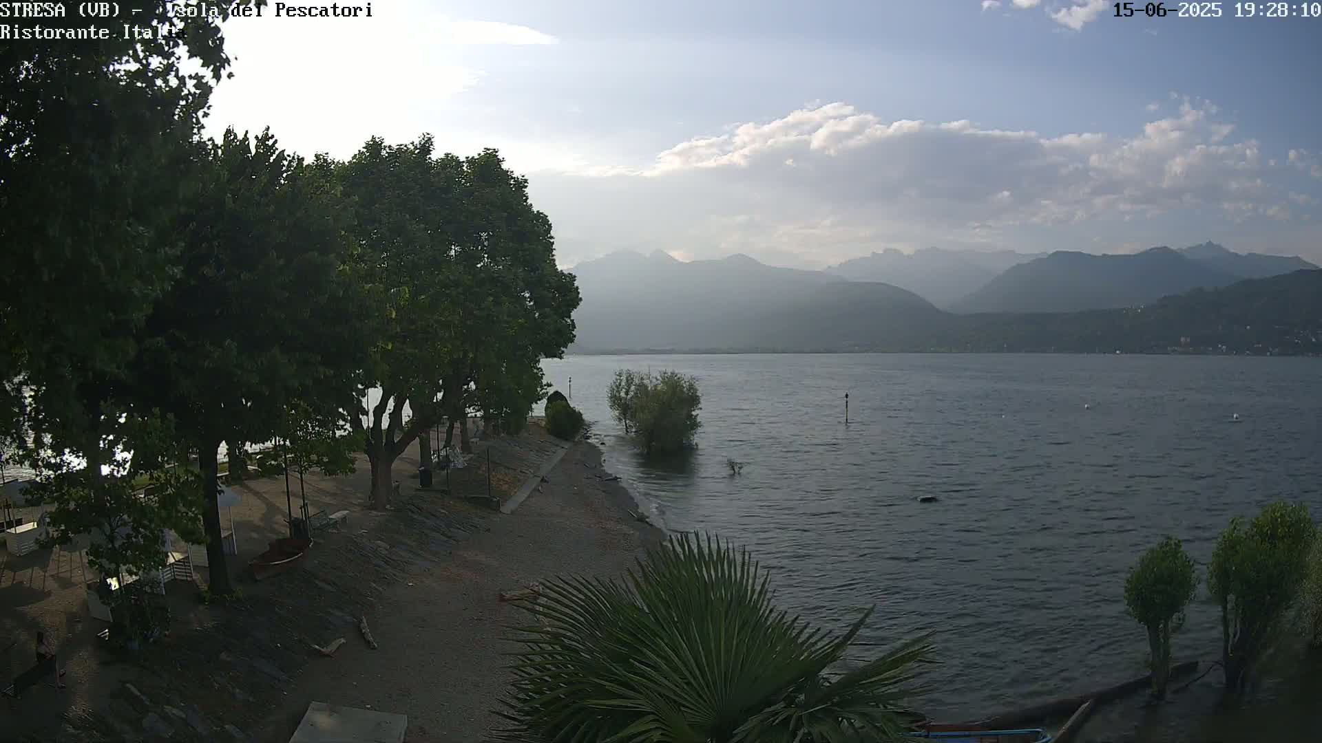 A partly cloudy evening scene shows a calm lake with mountains in the distance, a sandy shore with trees and some small structures, and a few people near the water.