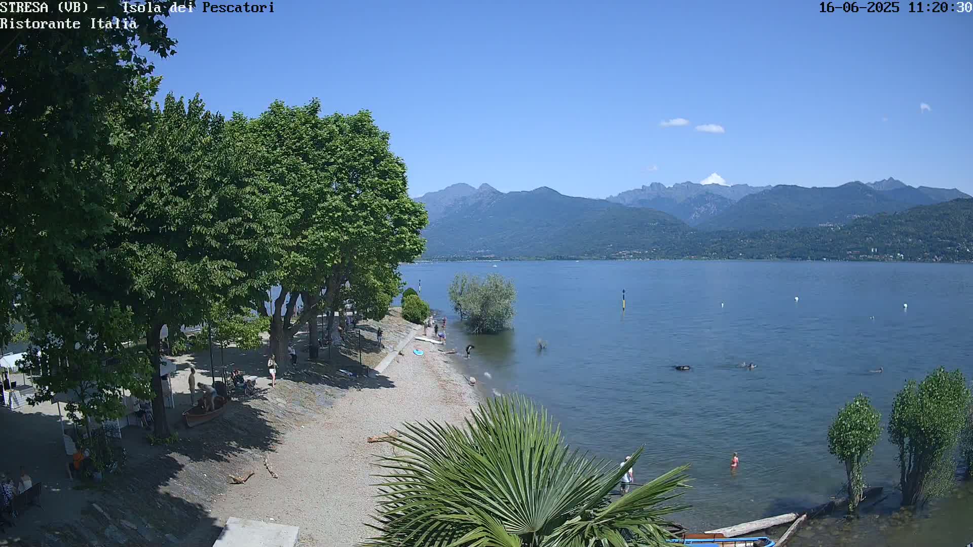 A sandy lakeshore with people swimming and relaxing under a clear blue sky, backed by lush green trees and distant mountains.