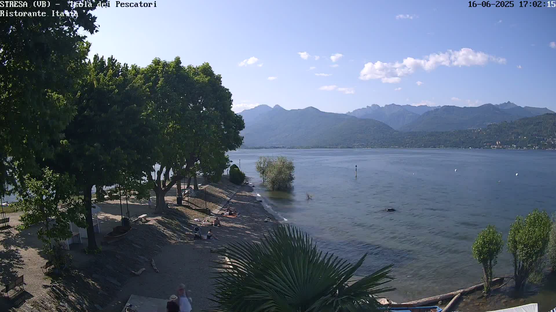 A sunny day shows a sandy beach with people relaxing under the shade of trees, overlooking a calm lake and distant mountains.