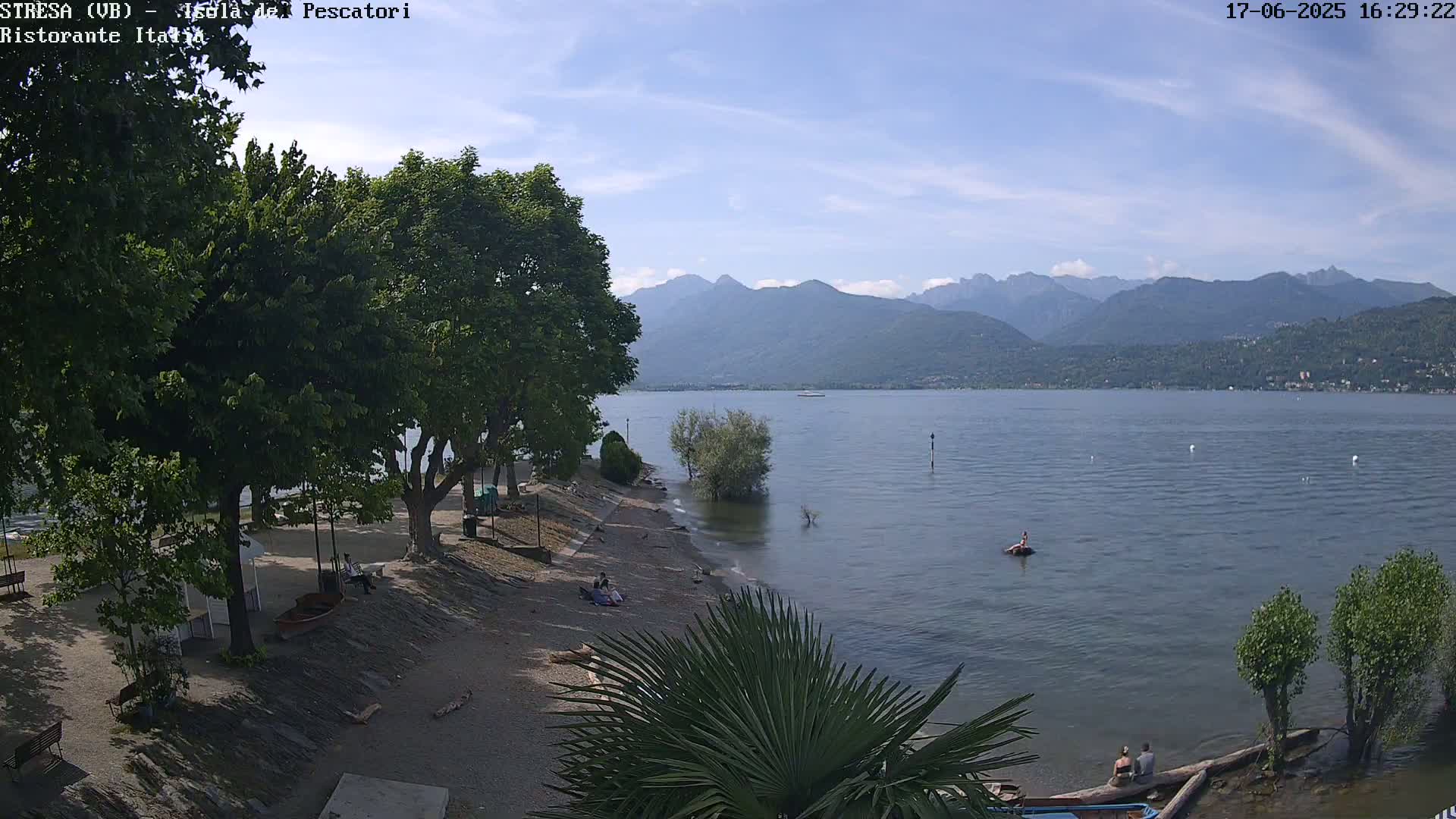 A lakeside beach scene on a partly sunny day shows a calm lake with mountains in the background, people relaxing on the beach and in the water, and lush green trees lining the shore.