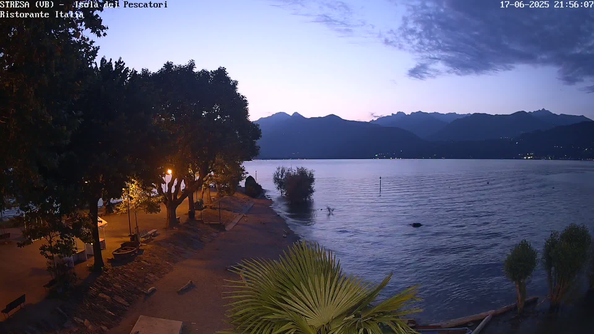A nighttime view of a calm lake with mountains in the distance, under a twilight sky, showing a shoreline with trees and lights.