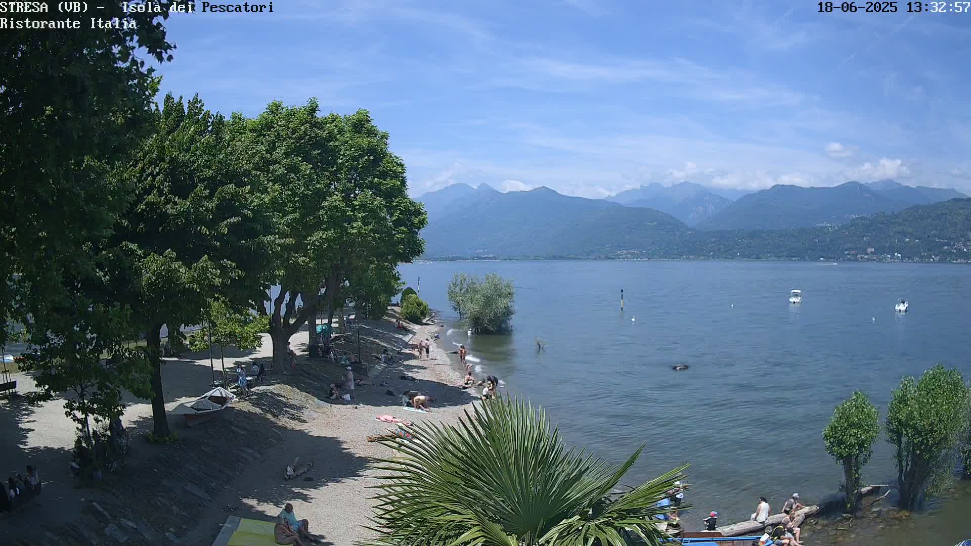 A sunny day at a lakeside beach shows people relaxing on a sandy shore, swimming in the calm water, and enjoying the view of distant mountains under a clear sky.