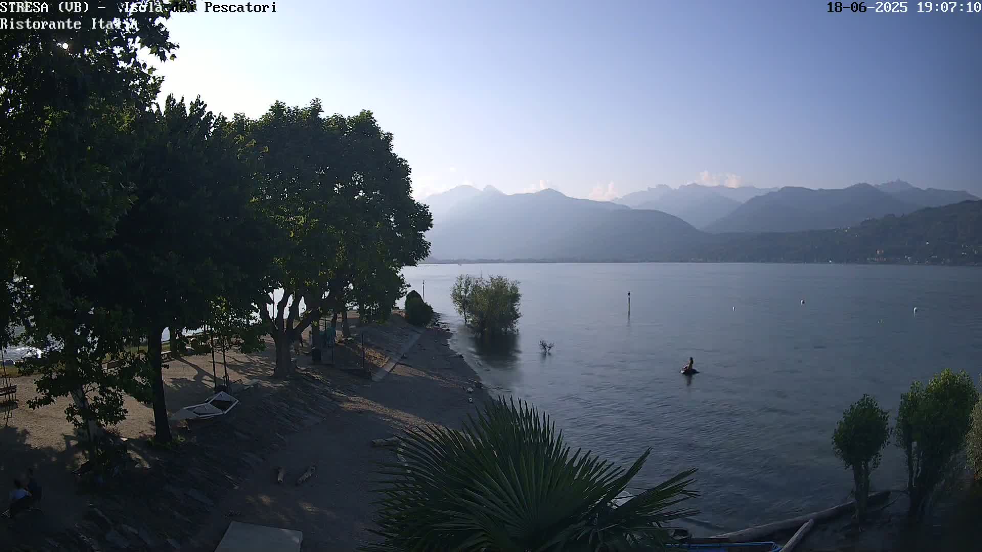 A calm lake under a clear, bright sky is seen from a lakeside vantage point with lush green trees and a sandy shore, mountains are visible in the background.