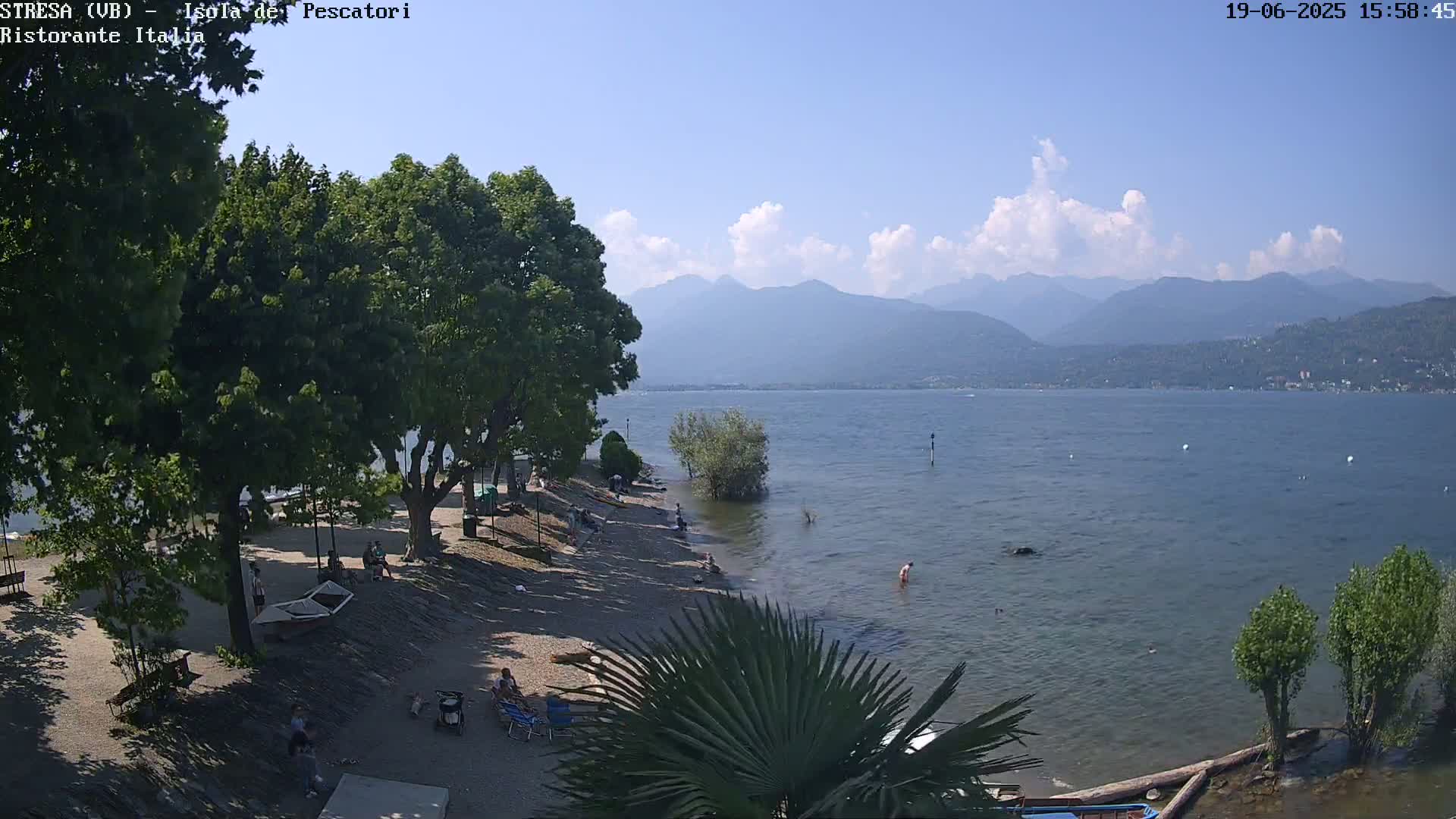 A sunny day at a lakeside beach with people relaxing, under partly cloudy skies and mountains in the distance.