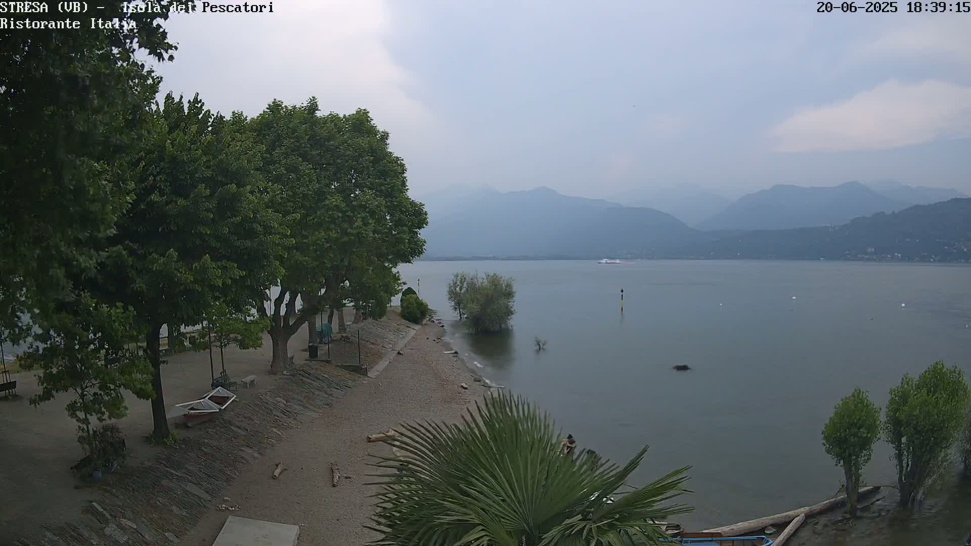 A calm lake with hazy mountains in the background, viewed from a lakeside area with trees and a small pebble beach under an overcast sky.
