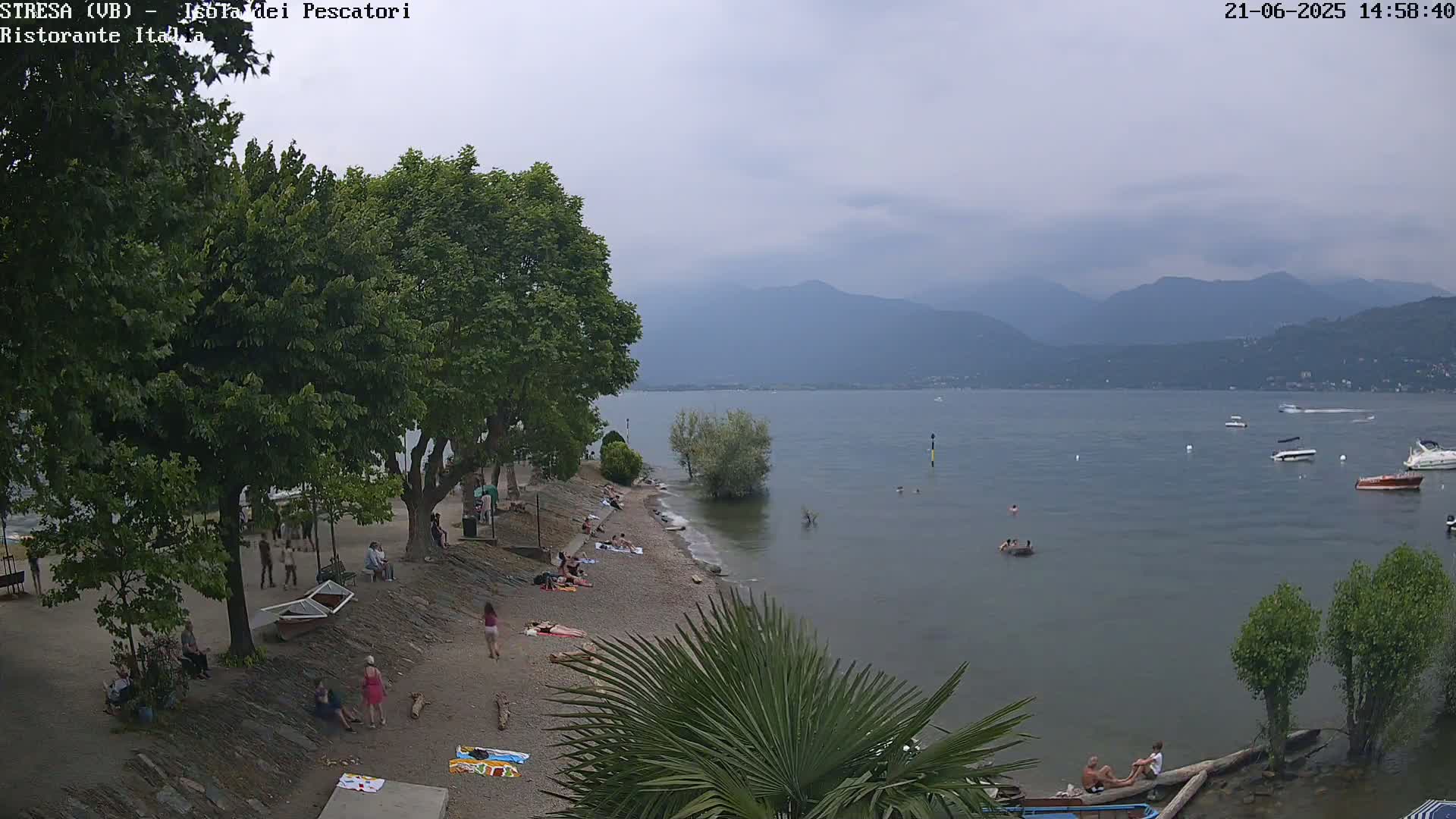 A pebble beach with people sunbathing and swimming in a calm lake, under an overcast sky, with mountains in the background.