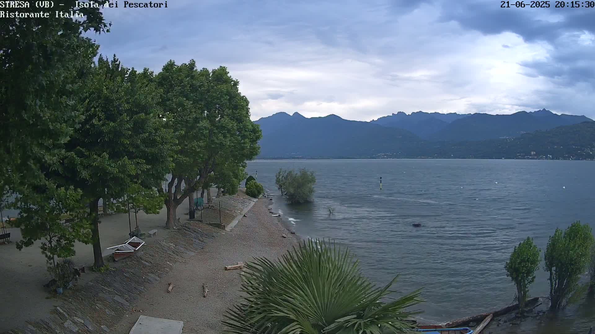 A mostly cloudy sky hangs over a calm lake, with mountains in the background and a pebble beach lined with trees and small boats in the foreground.