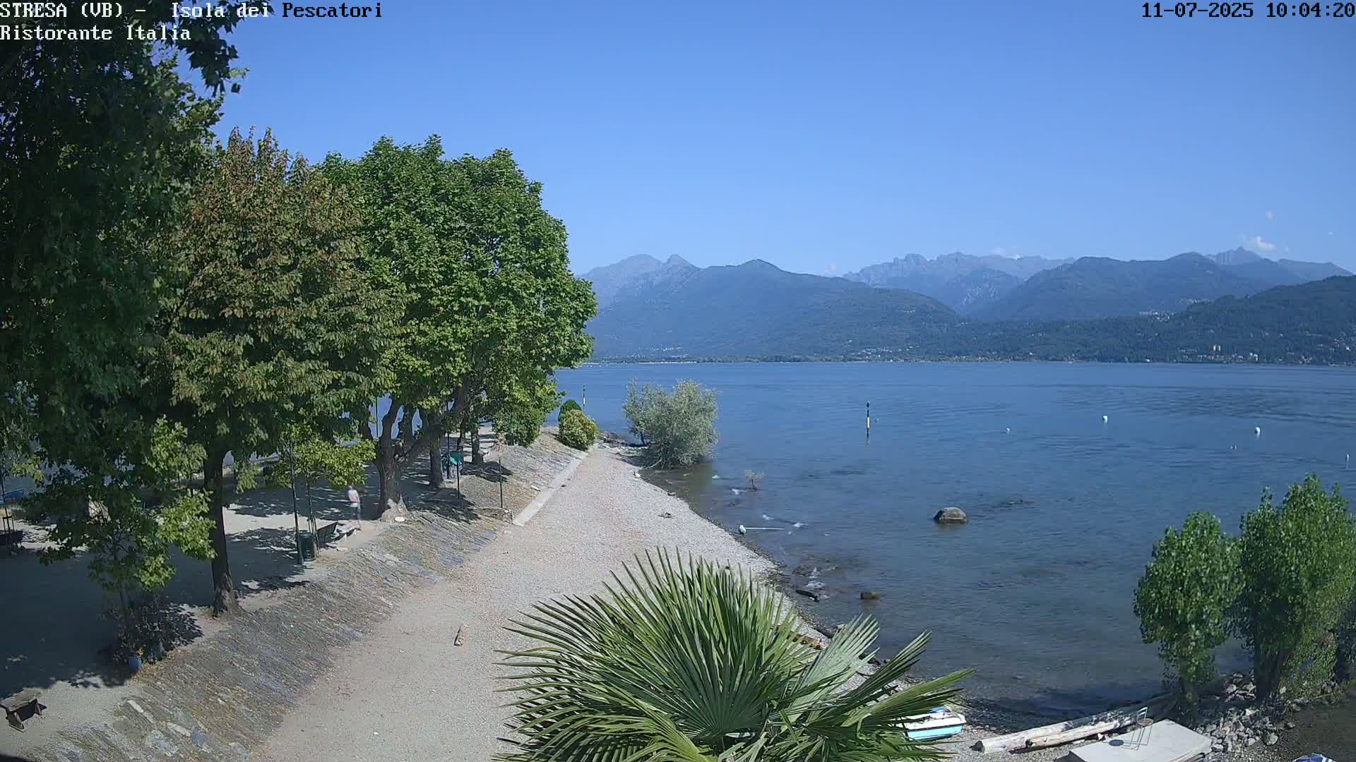 A pebble beach and tree-lined shore lead to a calm lake under a clear blue sky, with mountains in the distance.