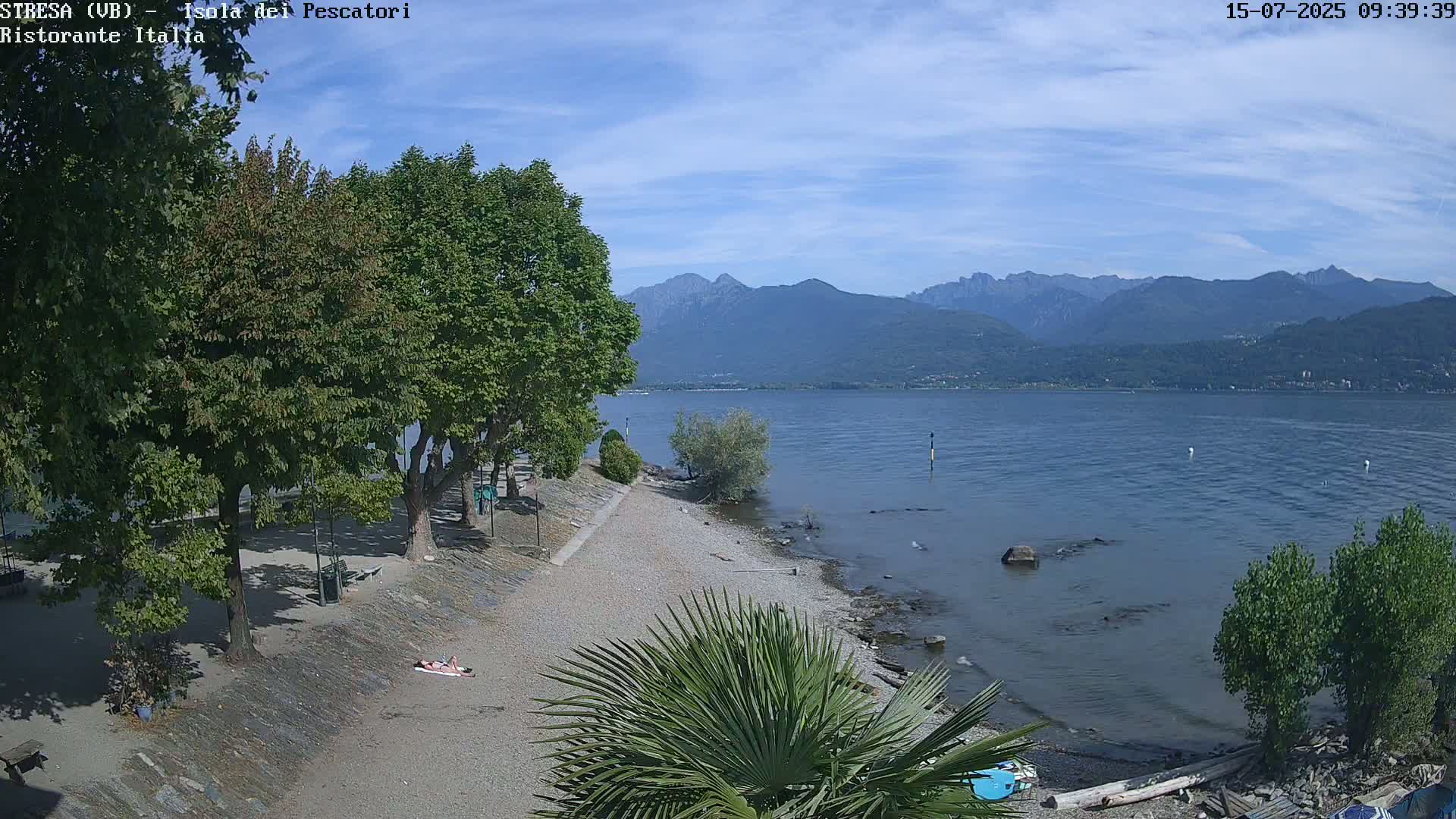 A person sunbathes on a pebble beach next to a calm lake, with a mountain range visible in the distance under a mostly clear, sunny sky.