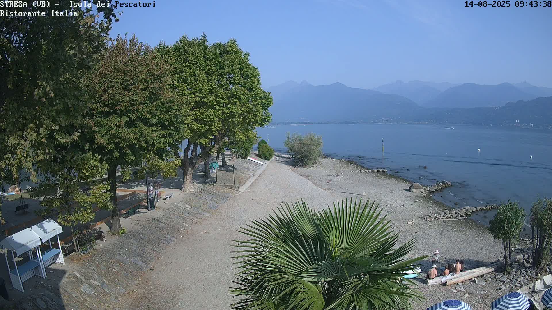 A pebble beach with people relaxing and several trees is shown, with calm water and mountains visible in the background under a clear sky.
