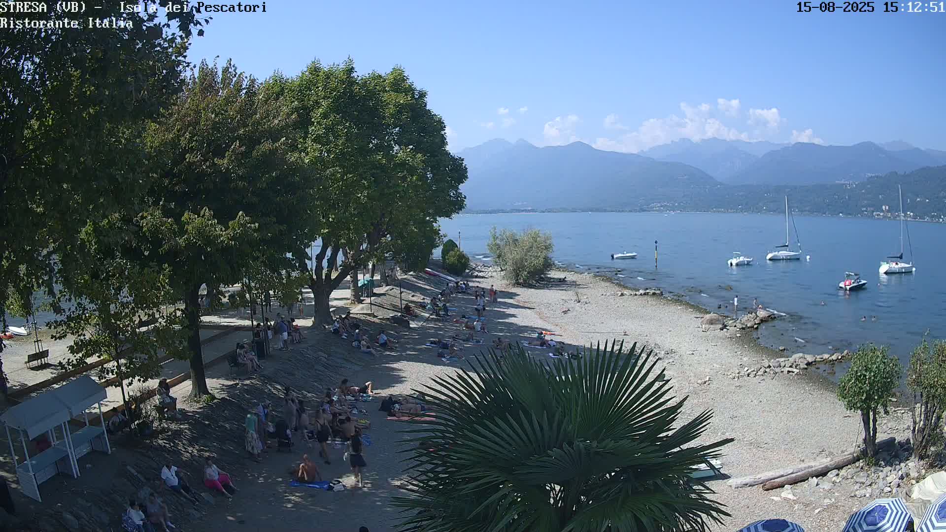 A pebble beach with people relaxing and several trees is shown, with calm water and mountains visible in the background under a clear sky.