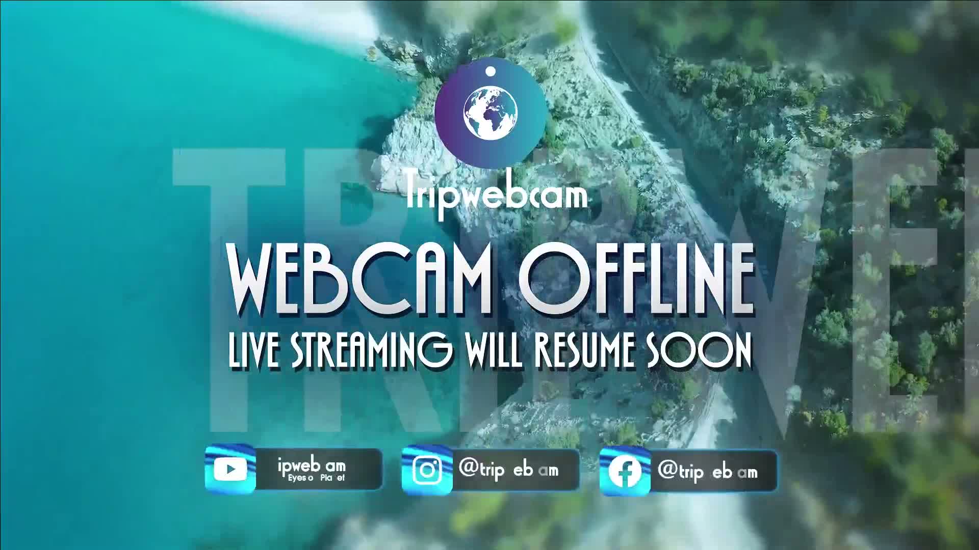 An aerial view shows a beautiful turquoise body of water bordered by green, tree-covered hills and a winding path, under clear skies.