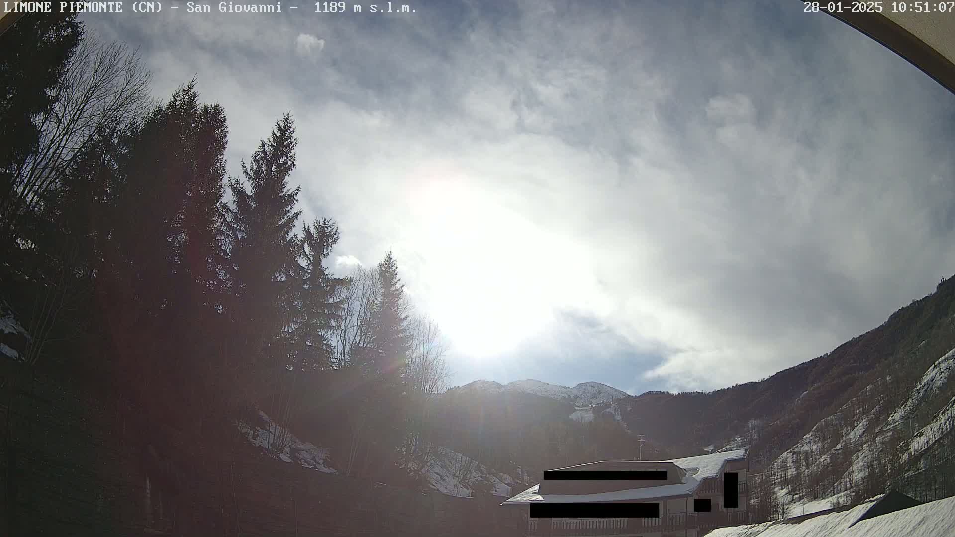 A partly cloudy, sunny day in a mountainous region shows a building partially obscured by evergreens and snow-dusted hills.
