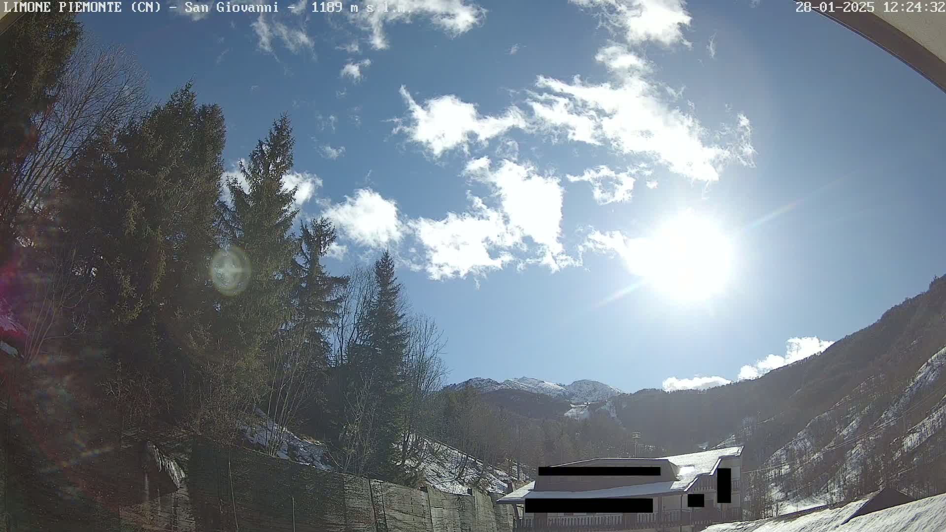 A sunny day with partly cloudy skies shows a snow-dusted mountainside and evergreen trees surrounding a building partially obscured by a fence.
