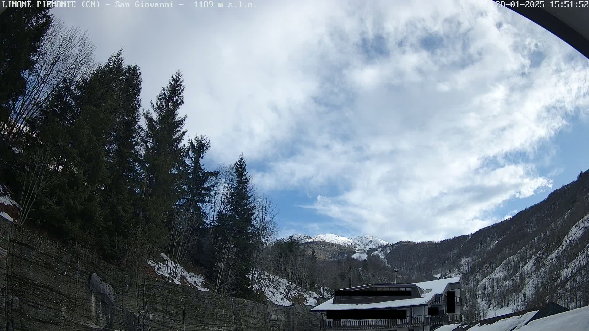 A partly cloudy day in a mountainous region shows evergreen trees, a snow-covered building, and snow on the ground and mountain slopes.
