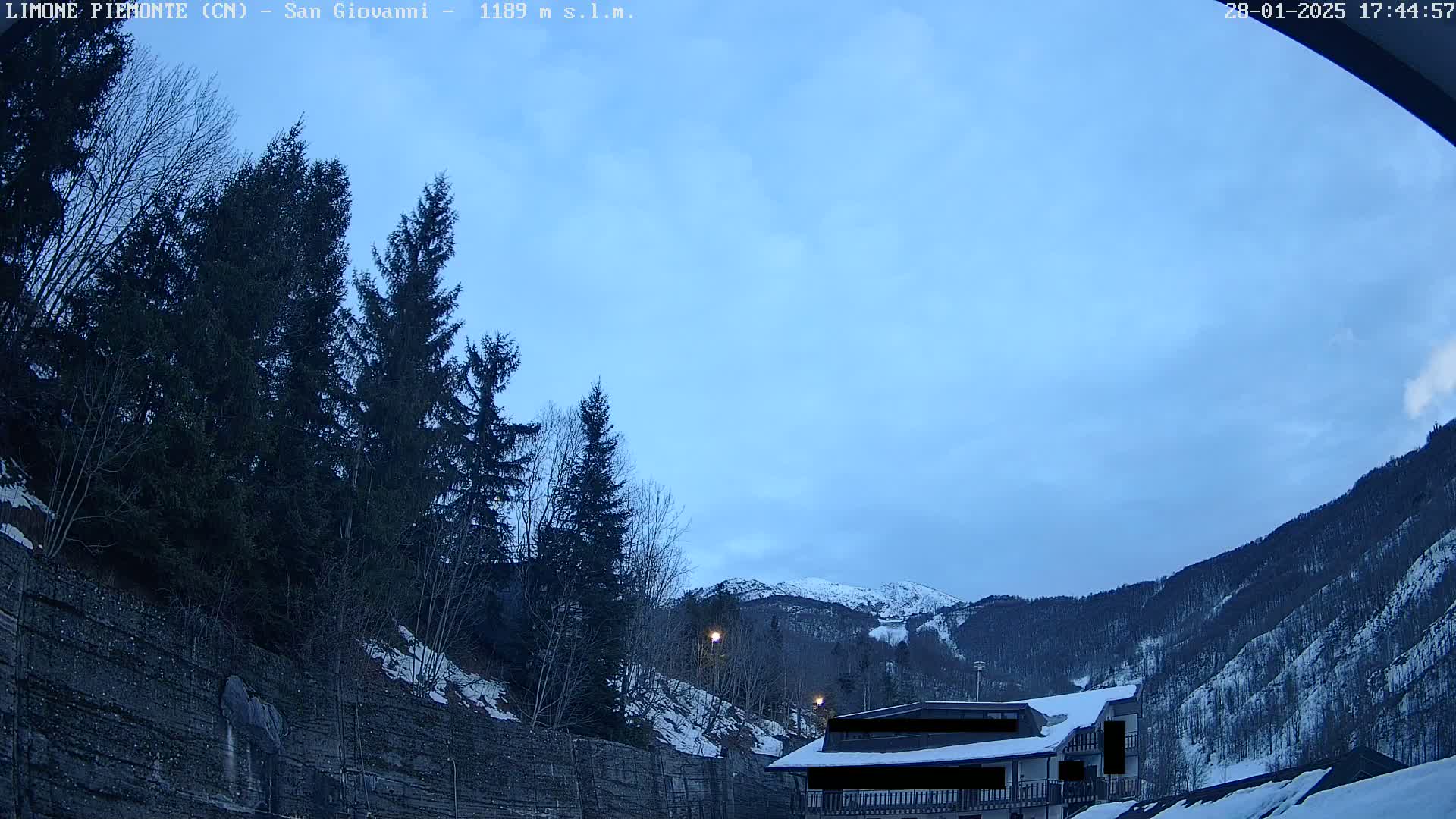 A partly cloudy twilight sky overlooks snow-dusted evergreen trees, a snow-covered hillside, and a building with a snow-covered roof.