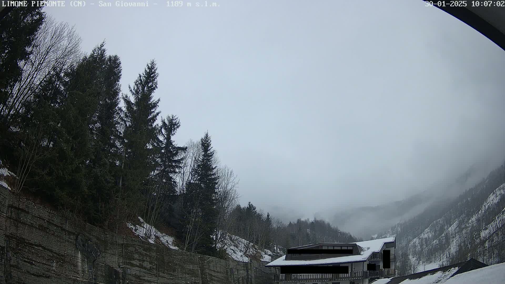 A foggy, overcast day reveals a mountainside with evergreens, a stone wall, and a building partially obscured by fog.