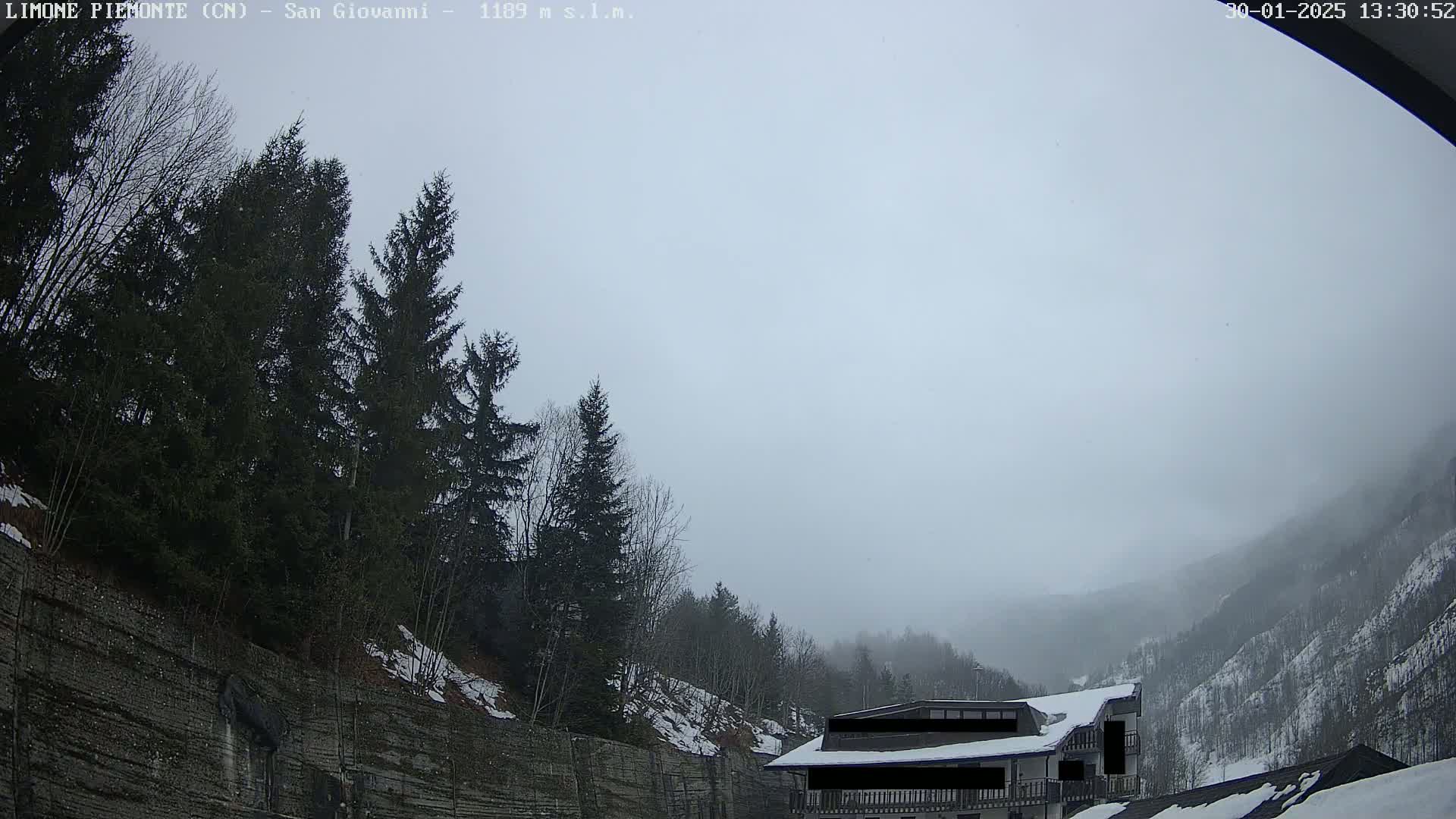 A foggy mountain valley scene shows evergreen trees and a partially obscured building with some snow on the ground.