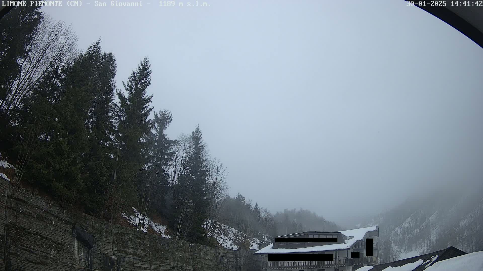 A foggy, snowy mountain scene shows evergreen trees, a stone wall, and a partially obscured building.