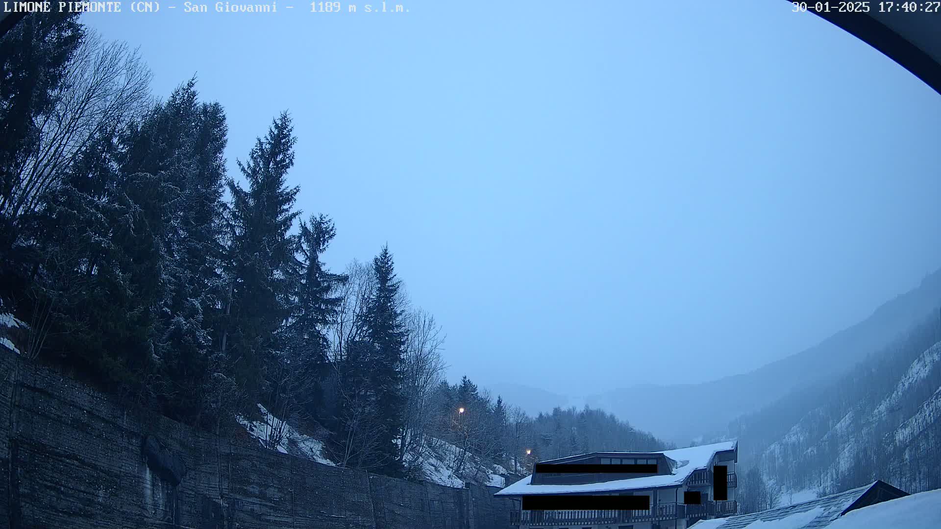 A hazy, light-blue sky hangs over snow-dusted evergreens and a mountain valley, partially obscured by a snow-covered building.
