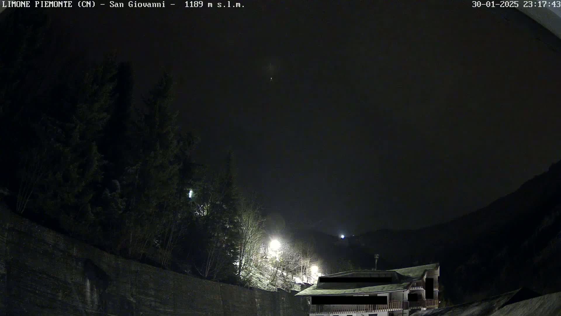 A nighttime view of a snow-dusted hillside with evergreens and a building, under a dark, clear sky.