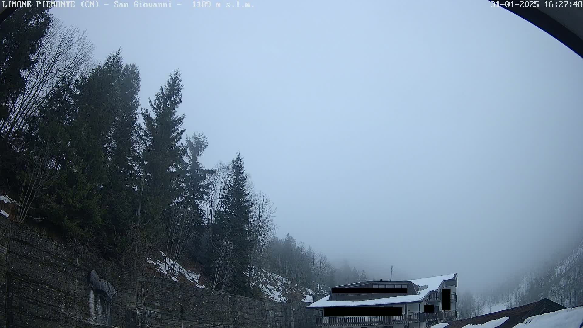 A foggy scene shows a building partially obscured by fog, nestled among evergreens on a snow-dusted hillside.