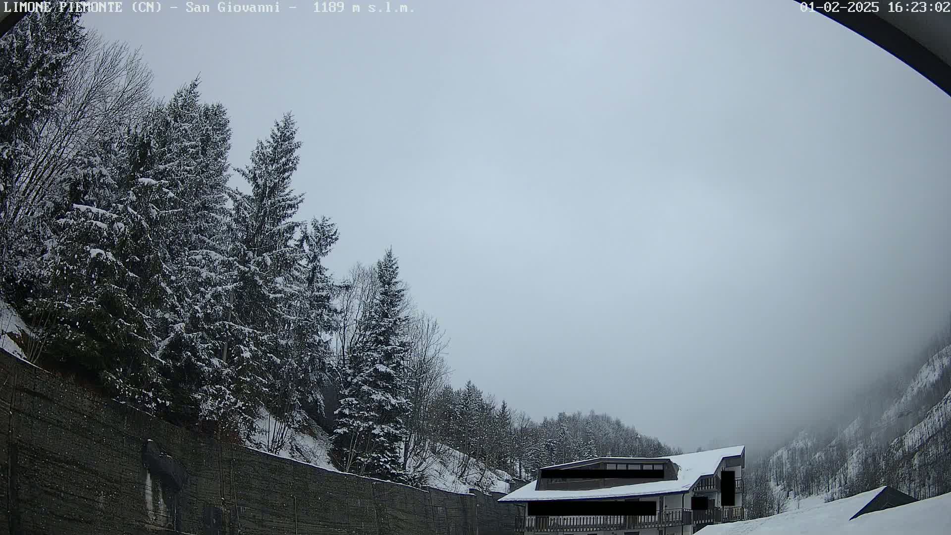 A snow-covered building sits in a valley surrounded by snow-covered evergreen trees under a hazy, overcast sky.