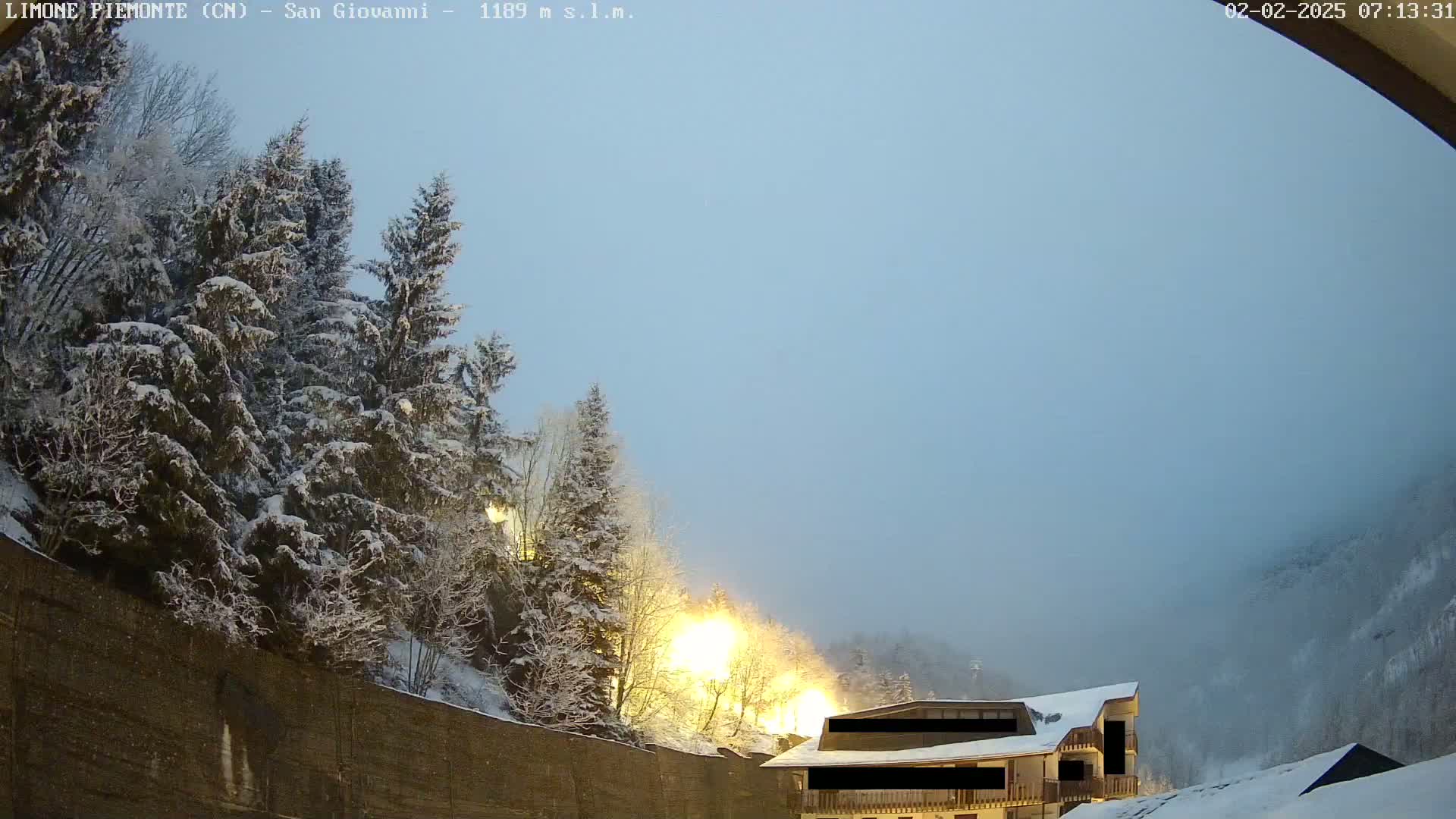 Snow-covered pine trees and a building are illuminated by lights under a hazy, light-blue sky.
