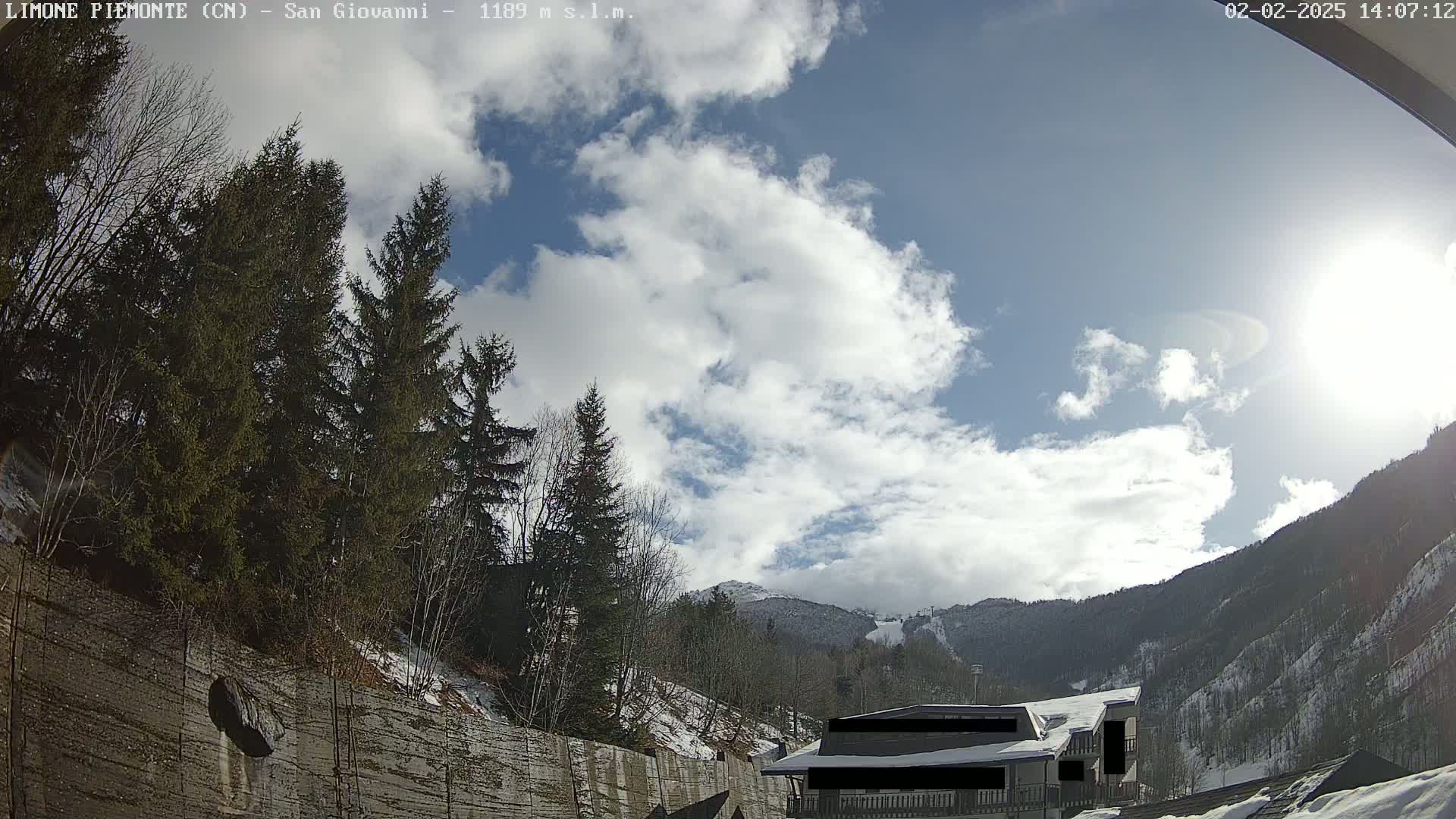 A partly sunny day with some clouds in a mountainous region shows evergreen trees and a building near a snow-covered hillside.