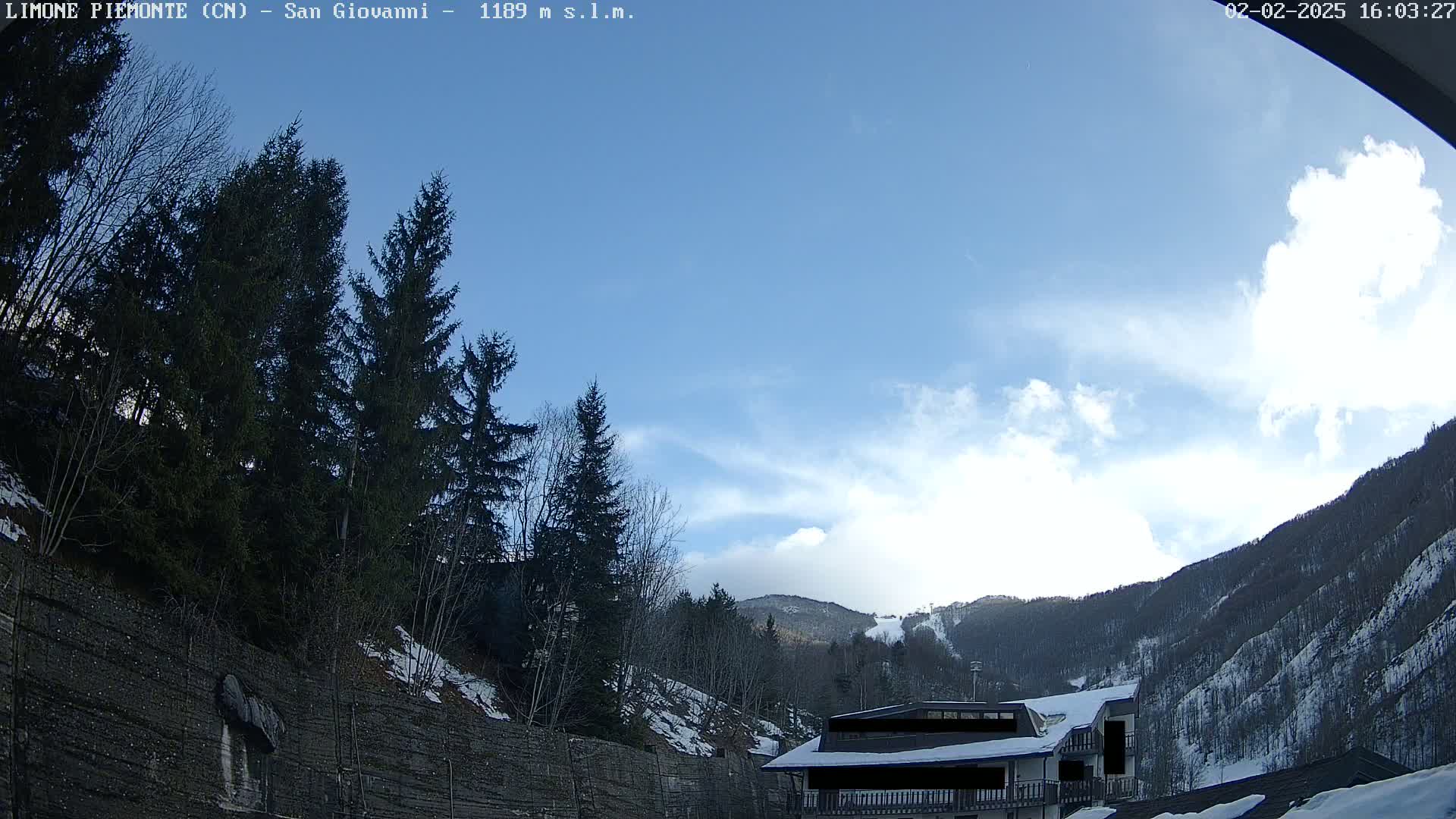 A mostly sunny day with some clouds in a mountainous region shows evergreen trees and a building with a snow-covered roof.