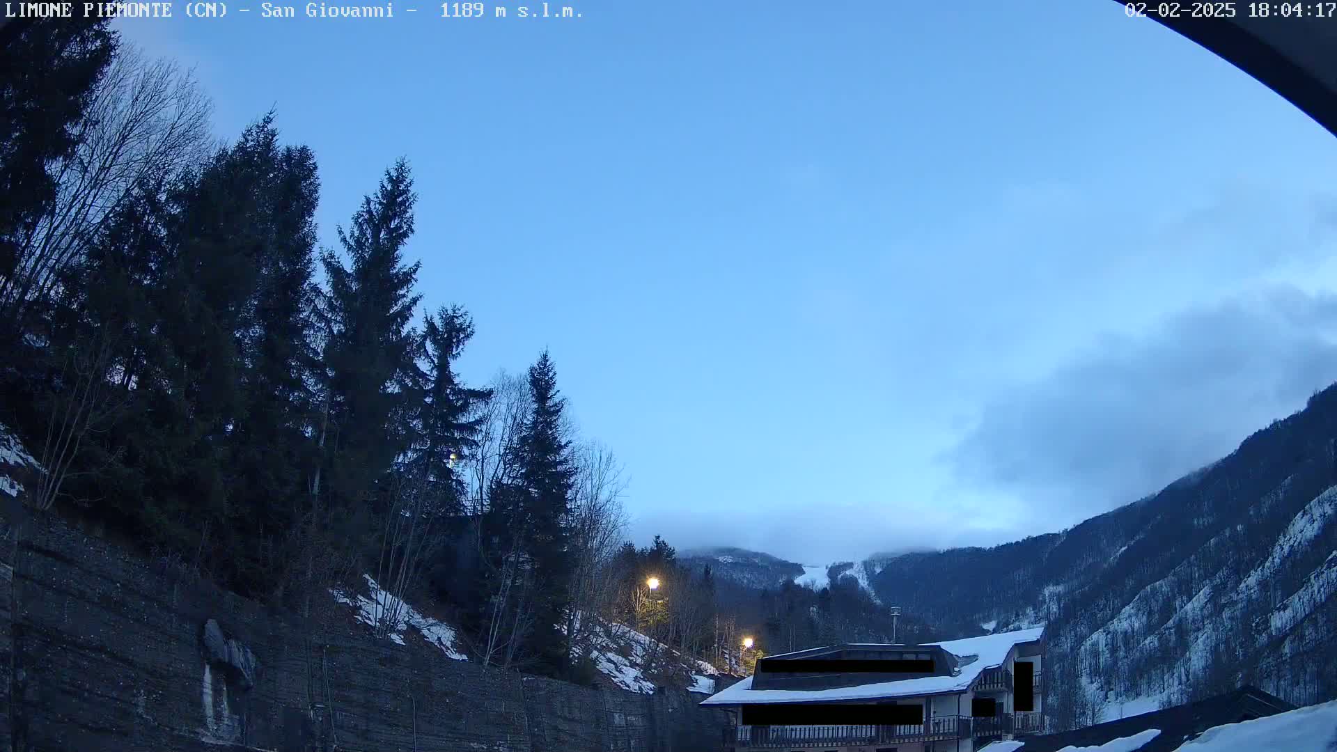 A twilight view of a mountainside with evergreen trees, a building, and some snow, under a mostly clear sky.