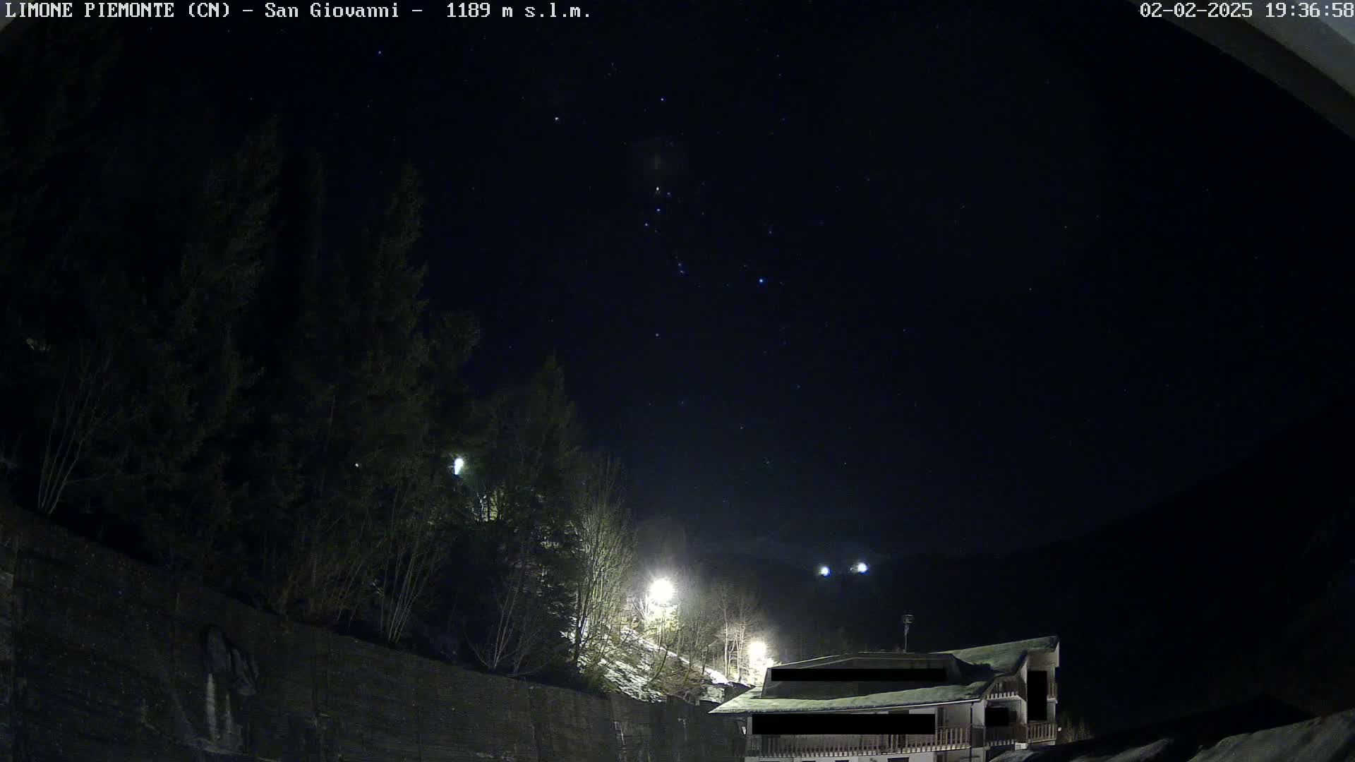A nighttime view of a snow-dusted hillside with trees and a building under a clear, starry sky.