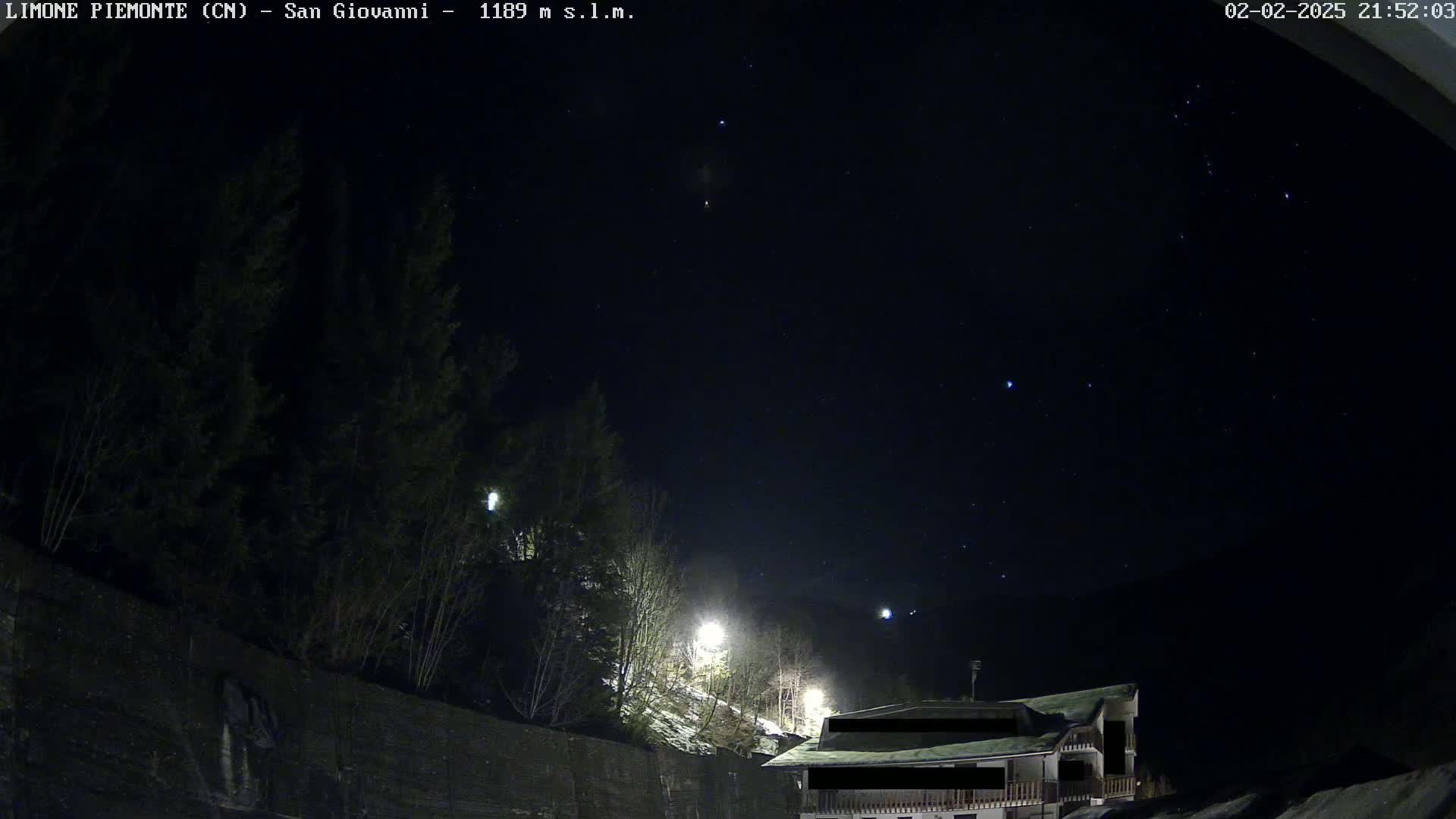 A nighttime view of a snow-covered hillside with trees and a building under a clear, starry sky.