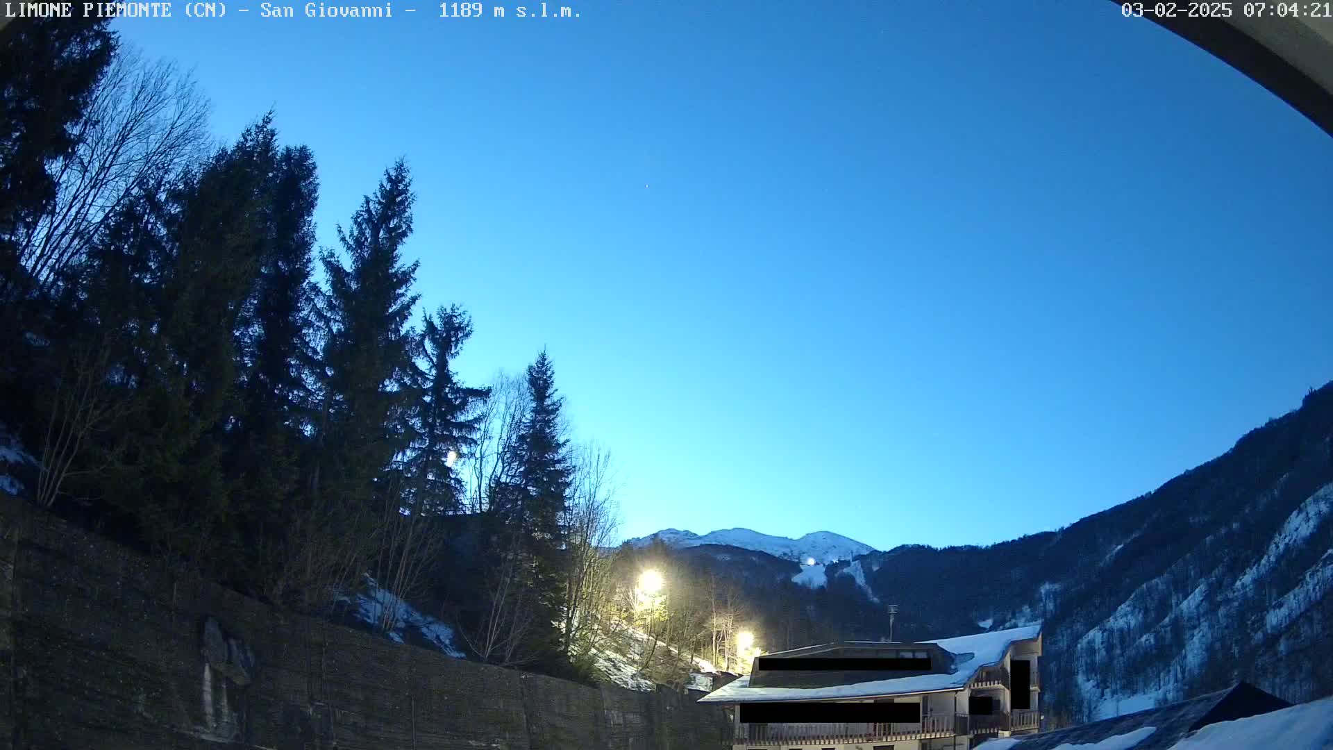Under a clear twilight sky, a building is illuminated near a snow-covered mountain and dark evergreen trees.