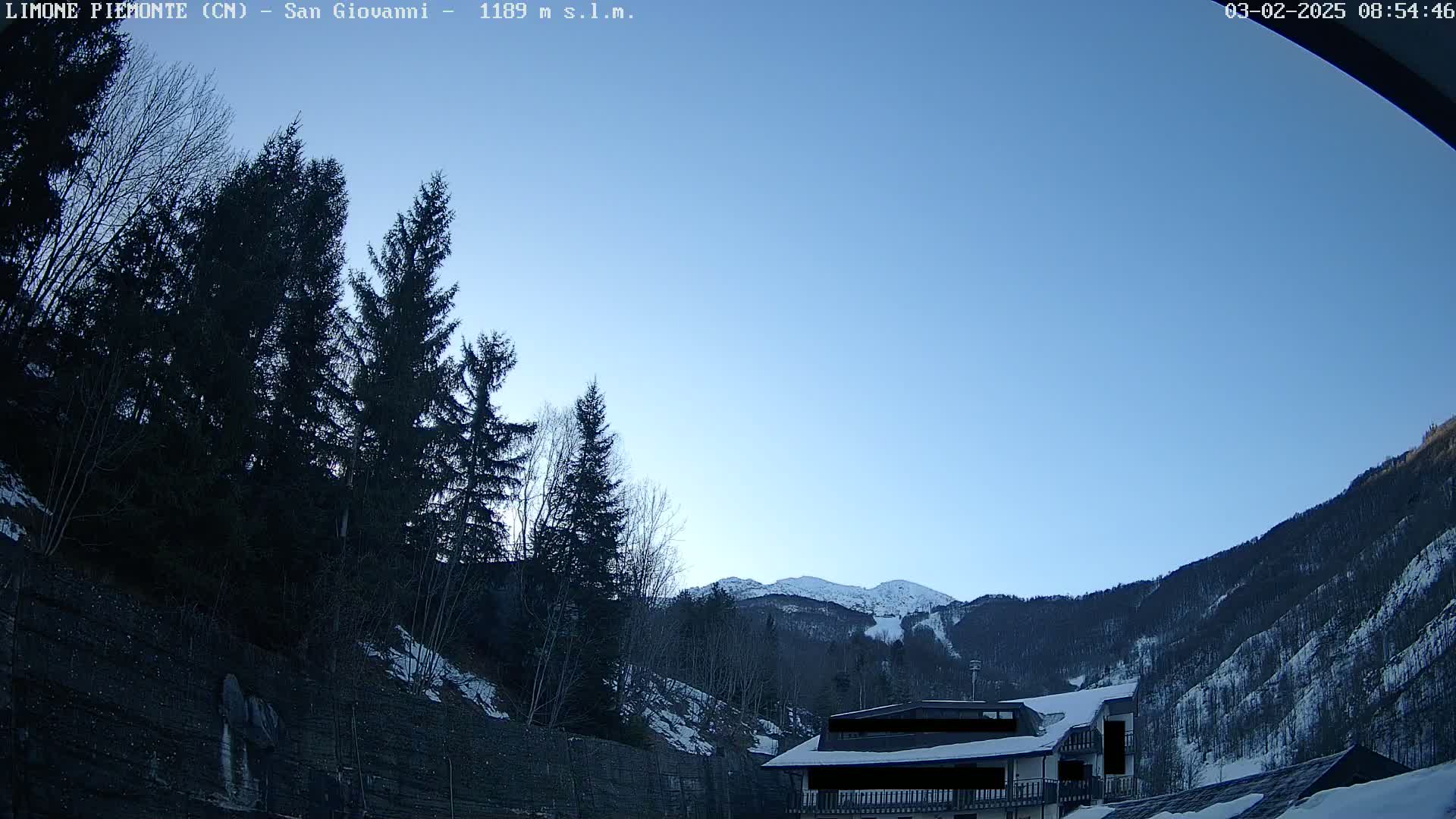A clear blue sky overlooks a snow-covered mountainside with dark evergreen trees and a building partially obscured in the foreground.