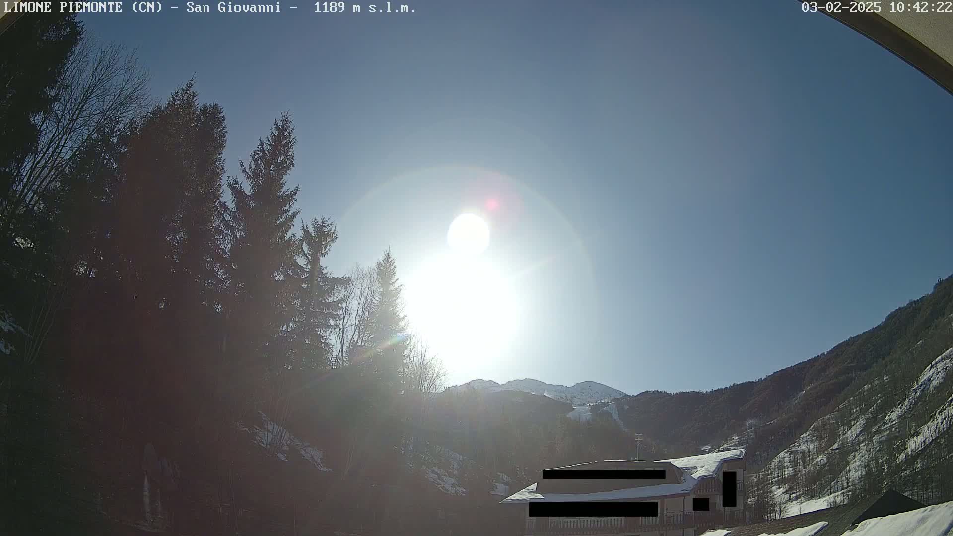 A bright, sunny day shows a mountainside with snow-covered trees and a building partially obscured by evergreens.