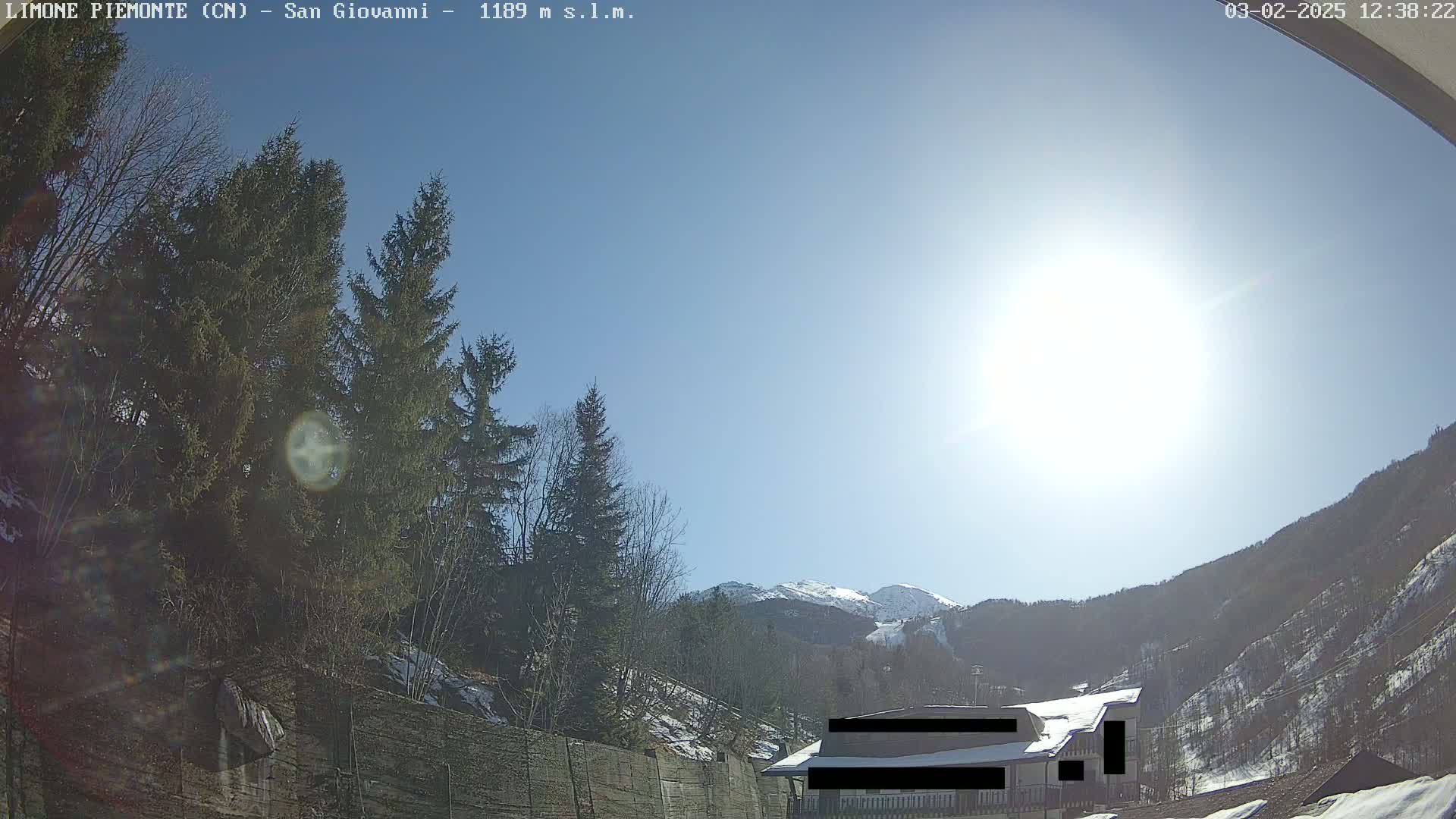 A sunny day in a mountainous region shows evergreen trees, a building partially obscured, and snow-capped mountains in the background.