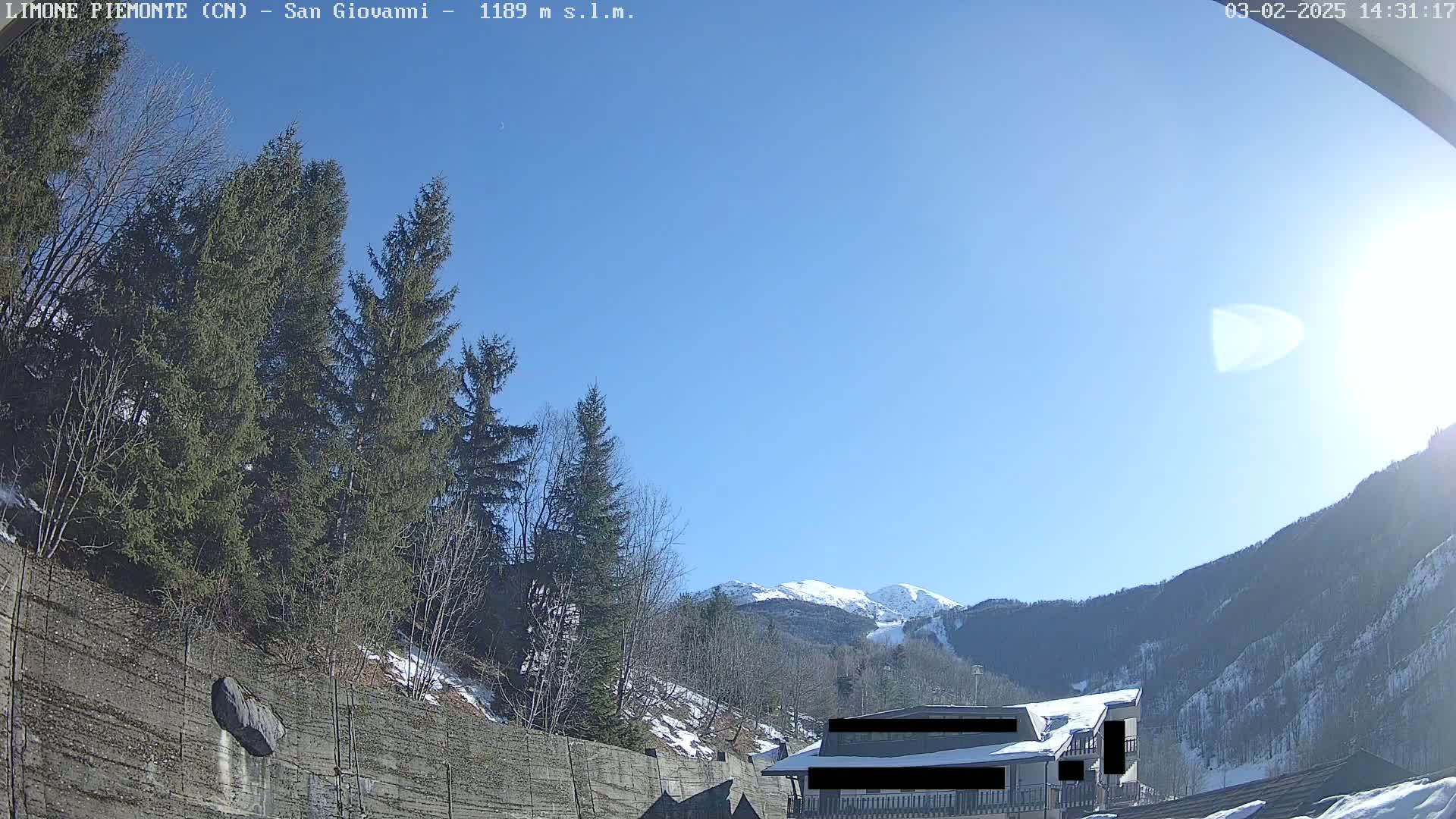 A sunny day in a mountainous region shows evergreen trees, a snow-covered hillside, and a building partially obscured by a retaining wall.
